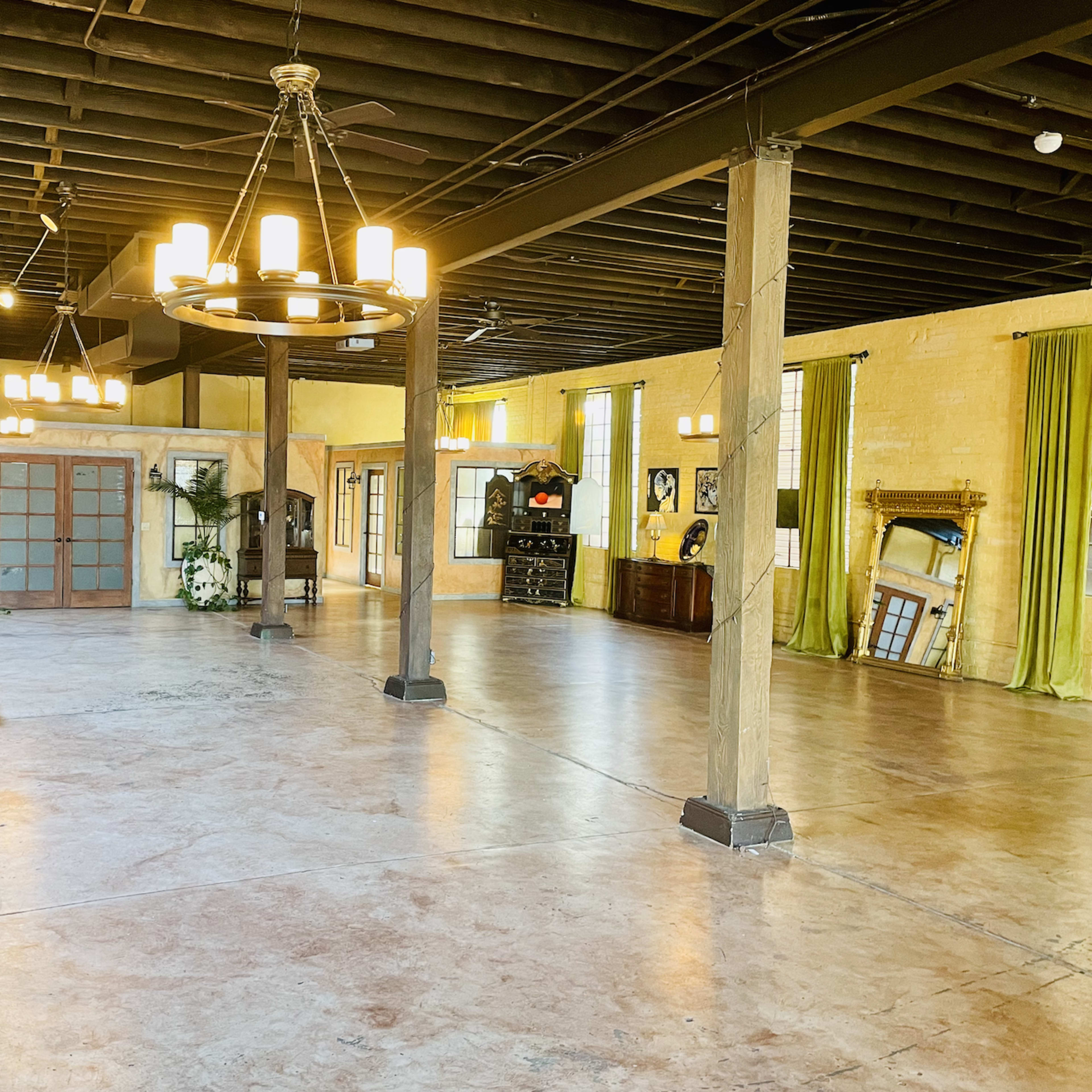 A large, open room with polished concrete floors, exposed beams, and chandeliers, featuring wooden furniture and decorative elements along the walls.