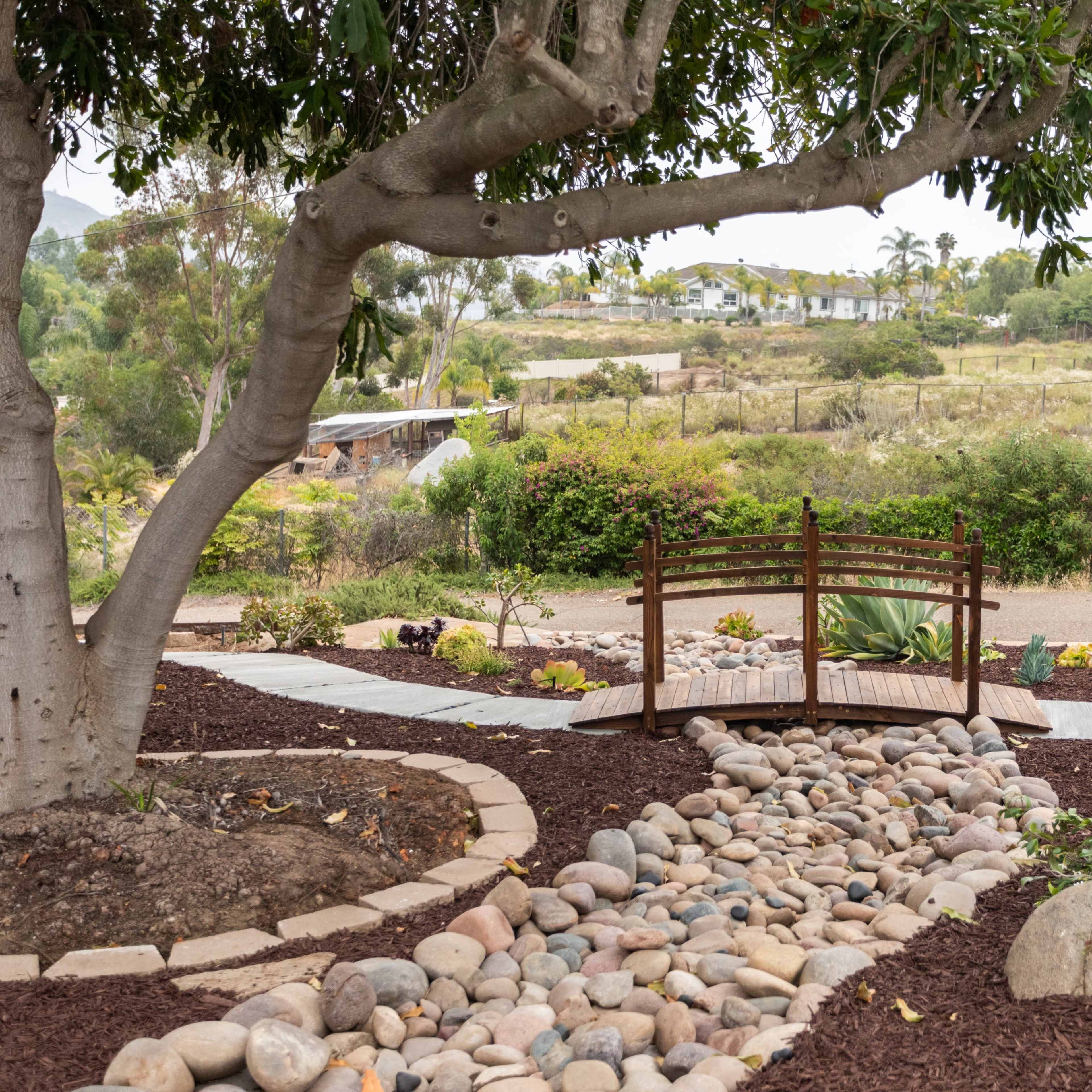 A wooden bridge spans over a riverbed of rocks in a landscaped garden with flower beds and a backdrop of greenery.