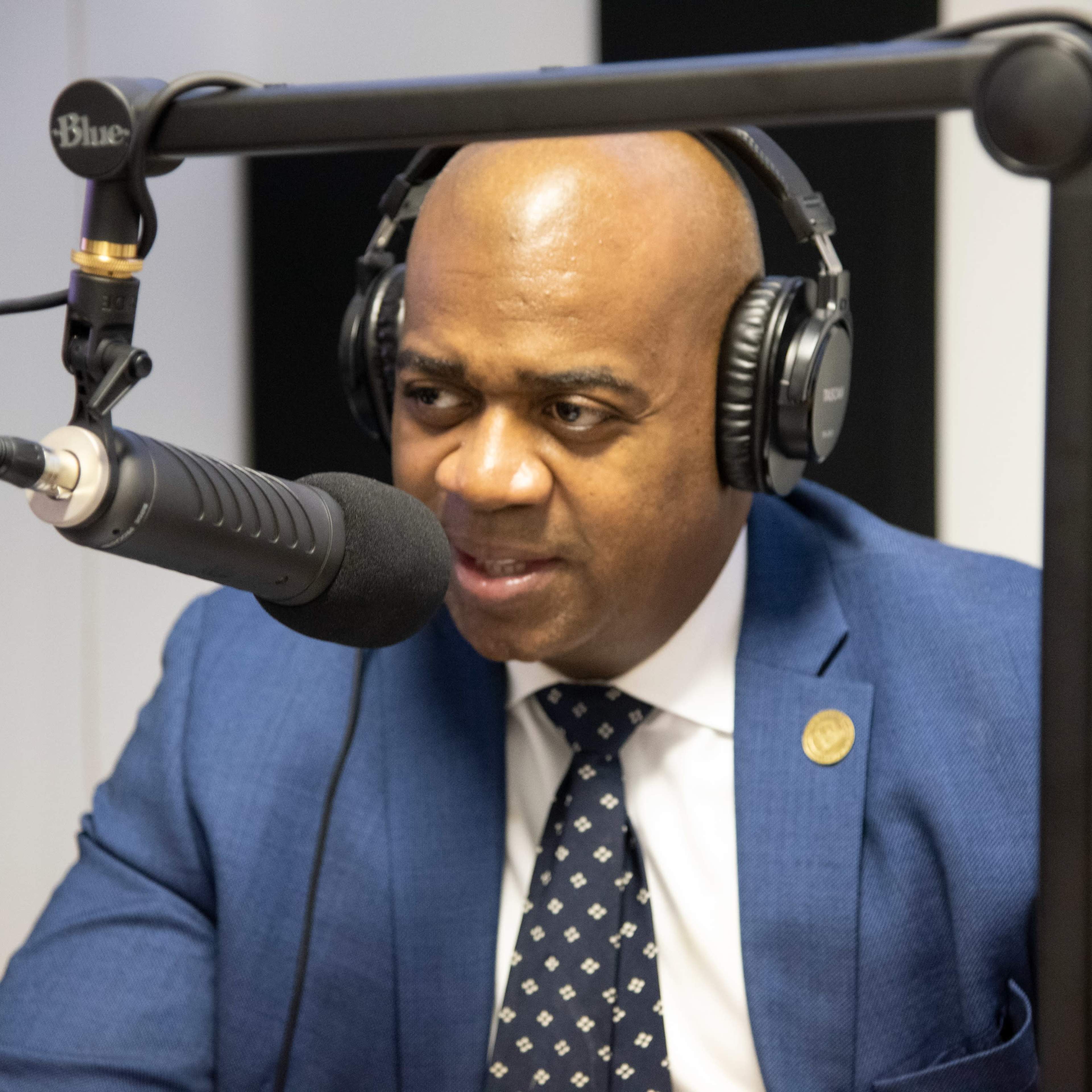 A man in a blue suit and headphones speaks into a microphone in a recording studio.