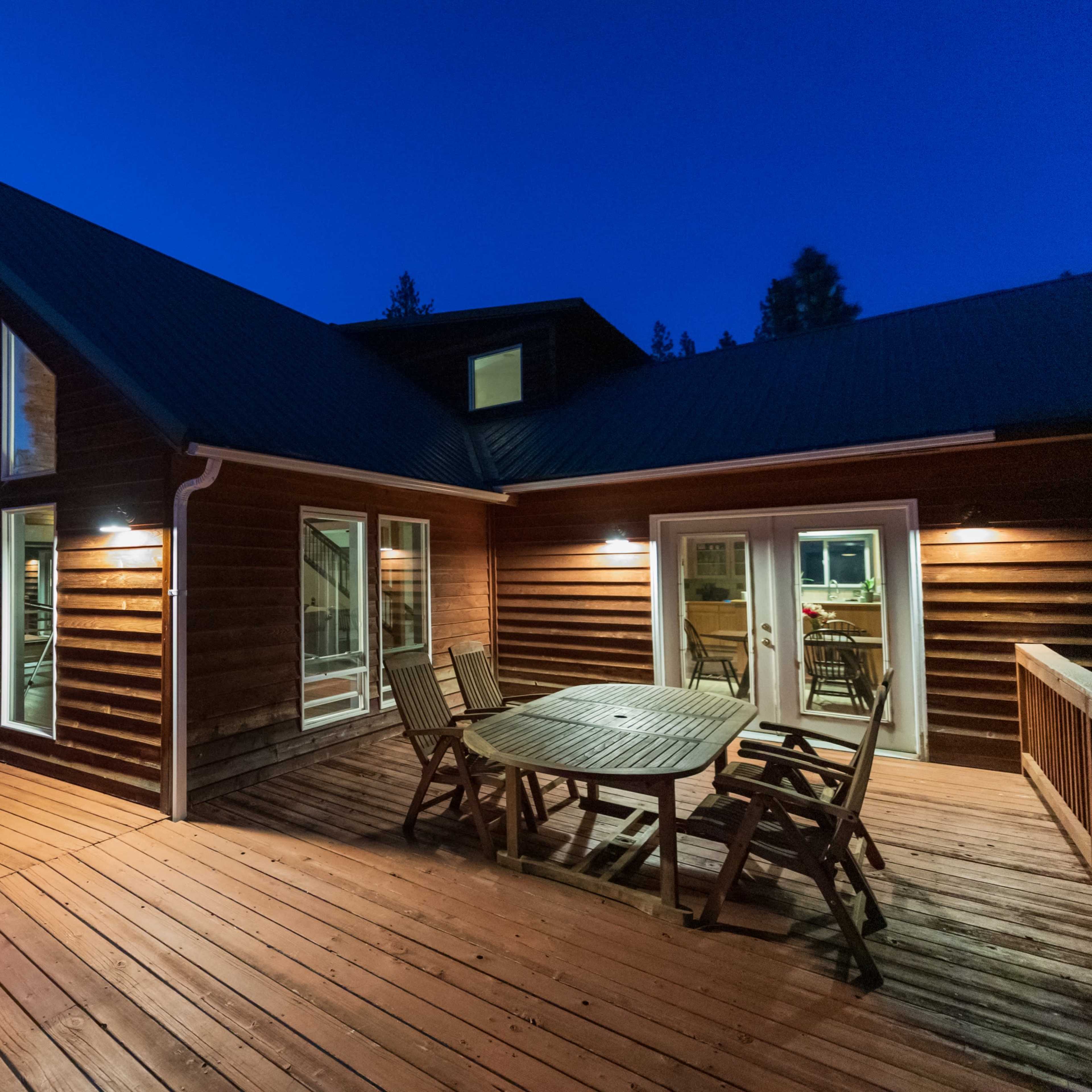 A wooden deck surrounds a modern house with large windows, situated in a wooded area at dusk.