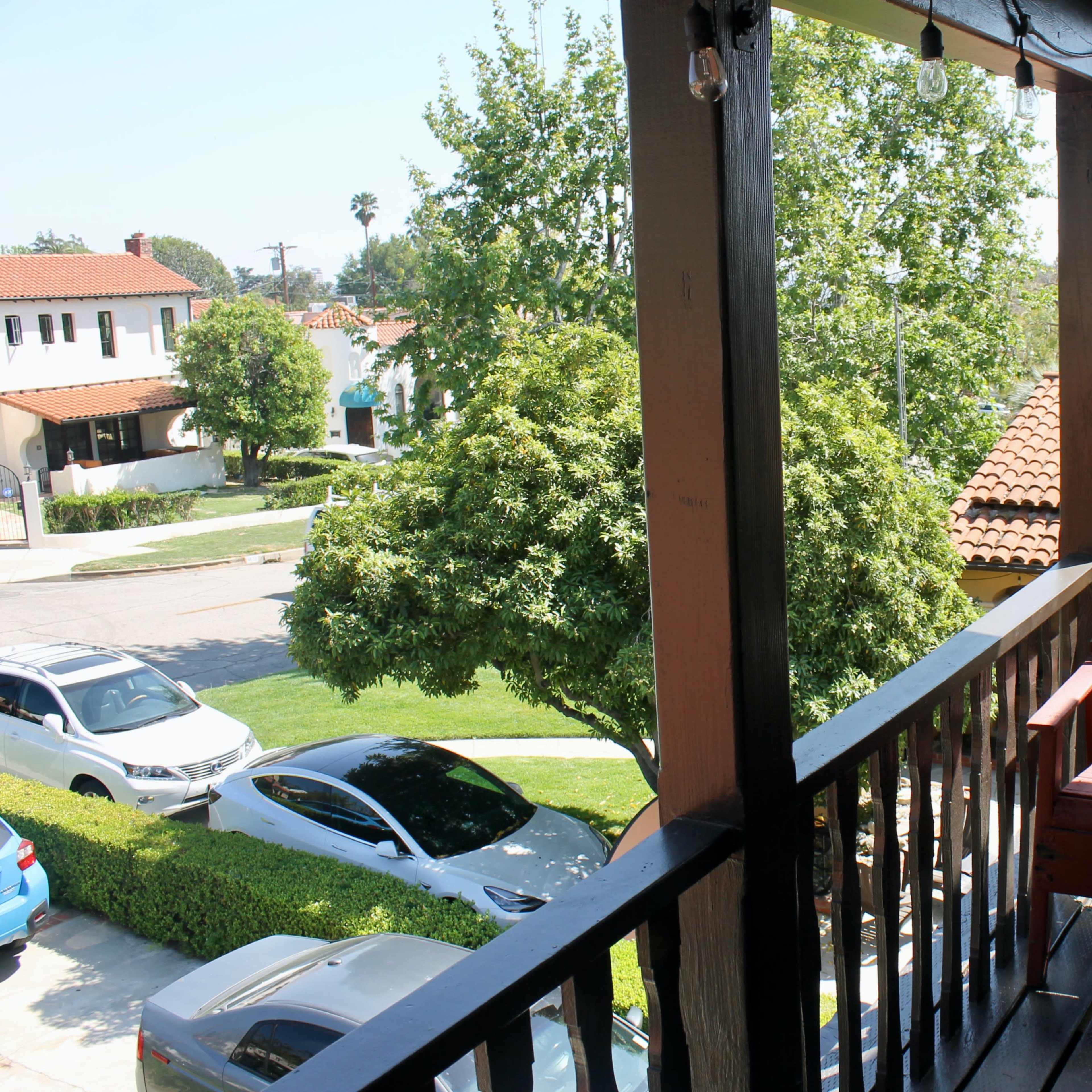 A balcony view overlooking a street with parked cars and houses, featuring a red adirondack chair and green trees in the foreground.