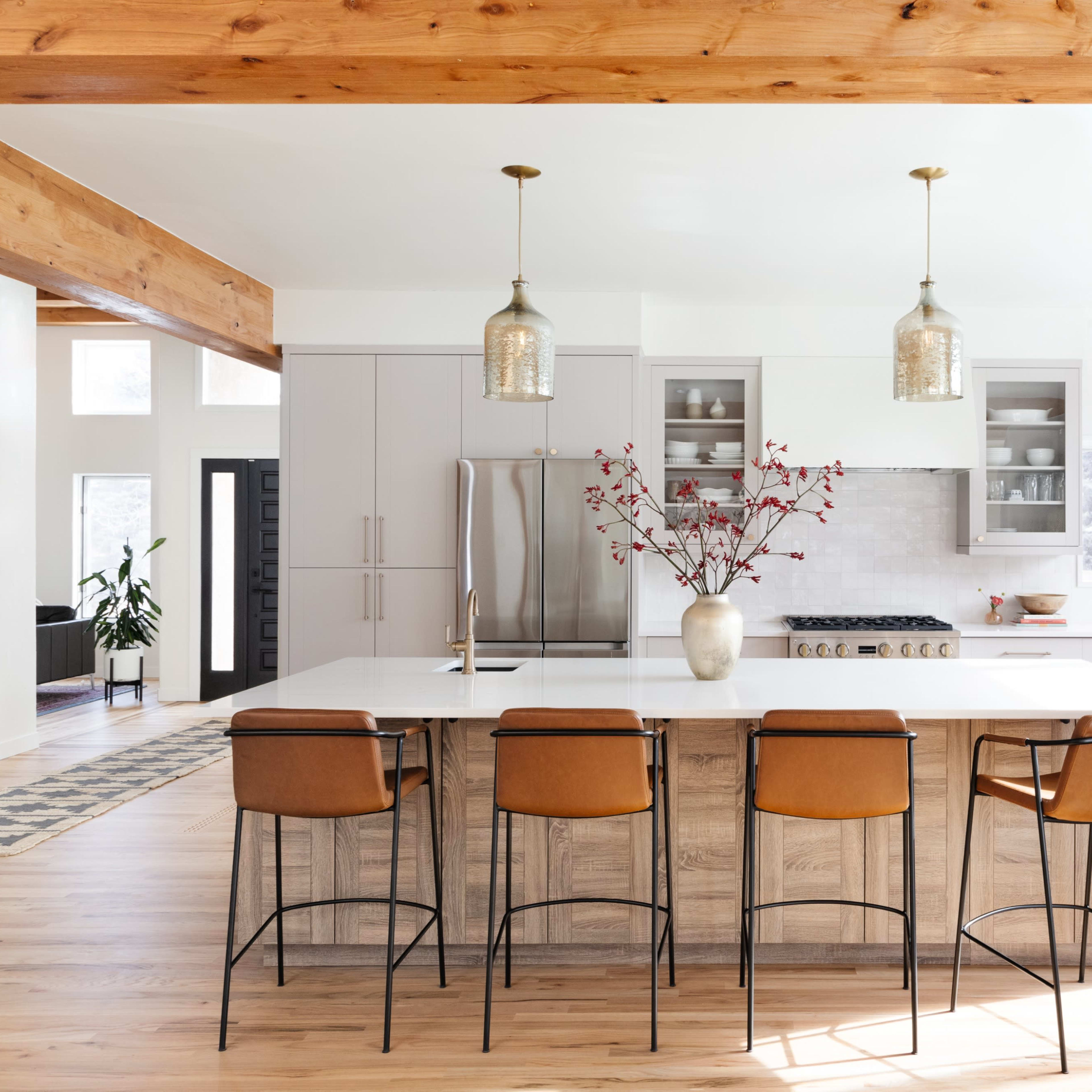 A modern kitchen features a central island with four leather bar stools, pendant lights, and a vase of branches, adjacent to an open living space with large windows.