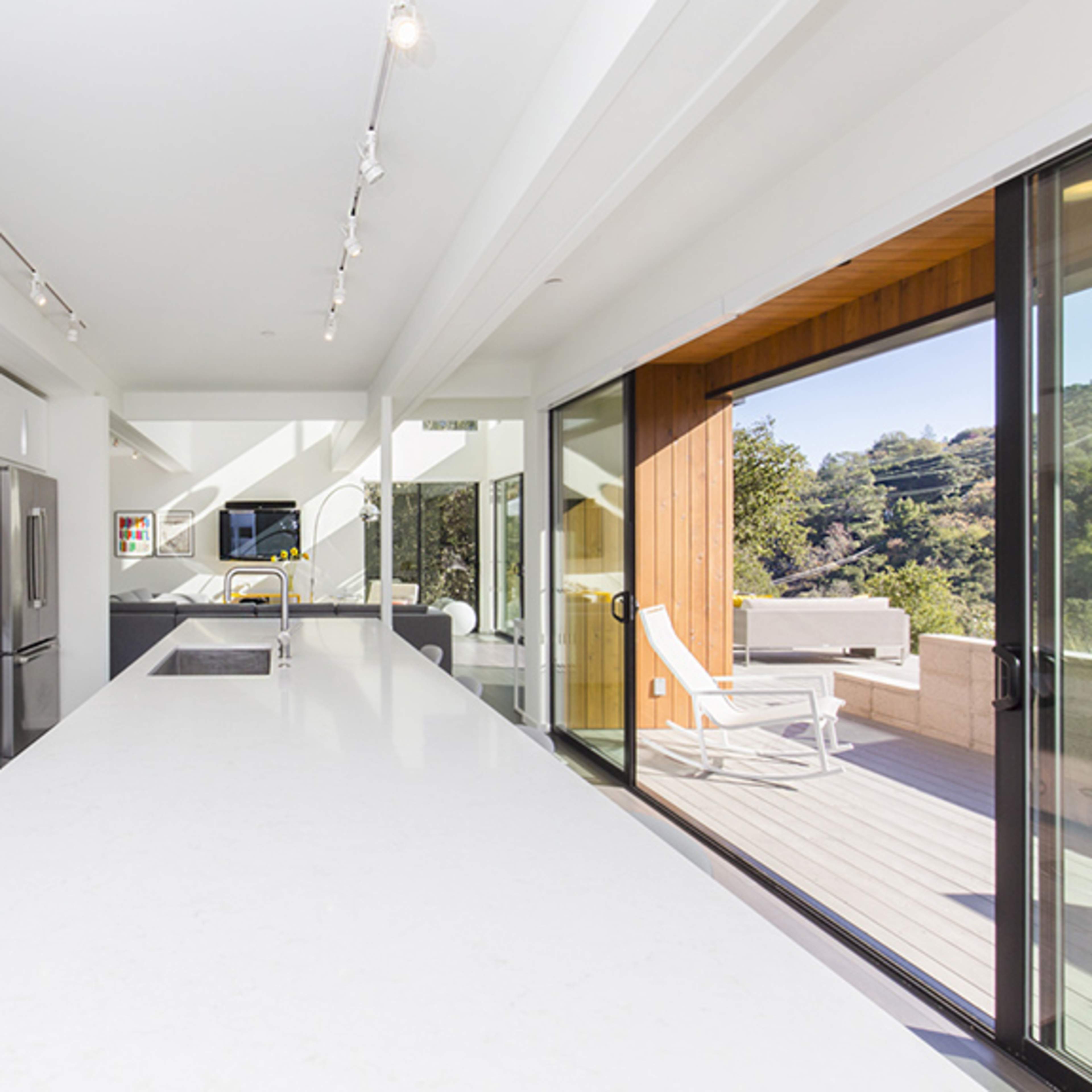 A modern kitchen with white cabinetry and an island, featuring large windows that offer a view of an outdoor deck.