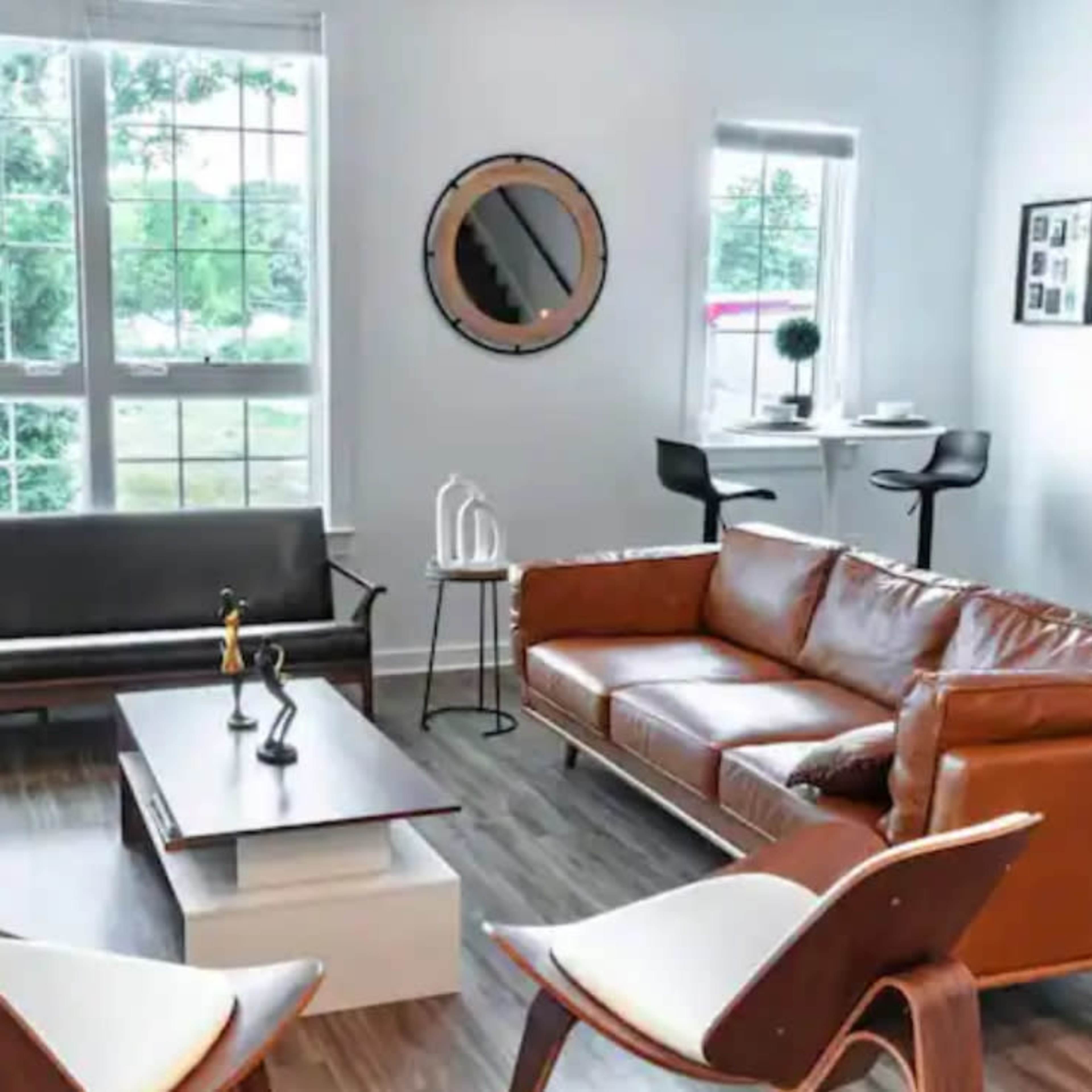 A modern living room with a leather sofa, a wooden coffee table, two armchairs, and large windows letting in natural light.