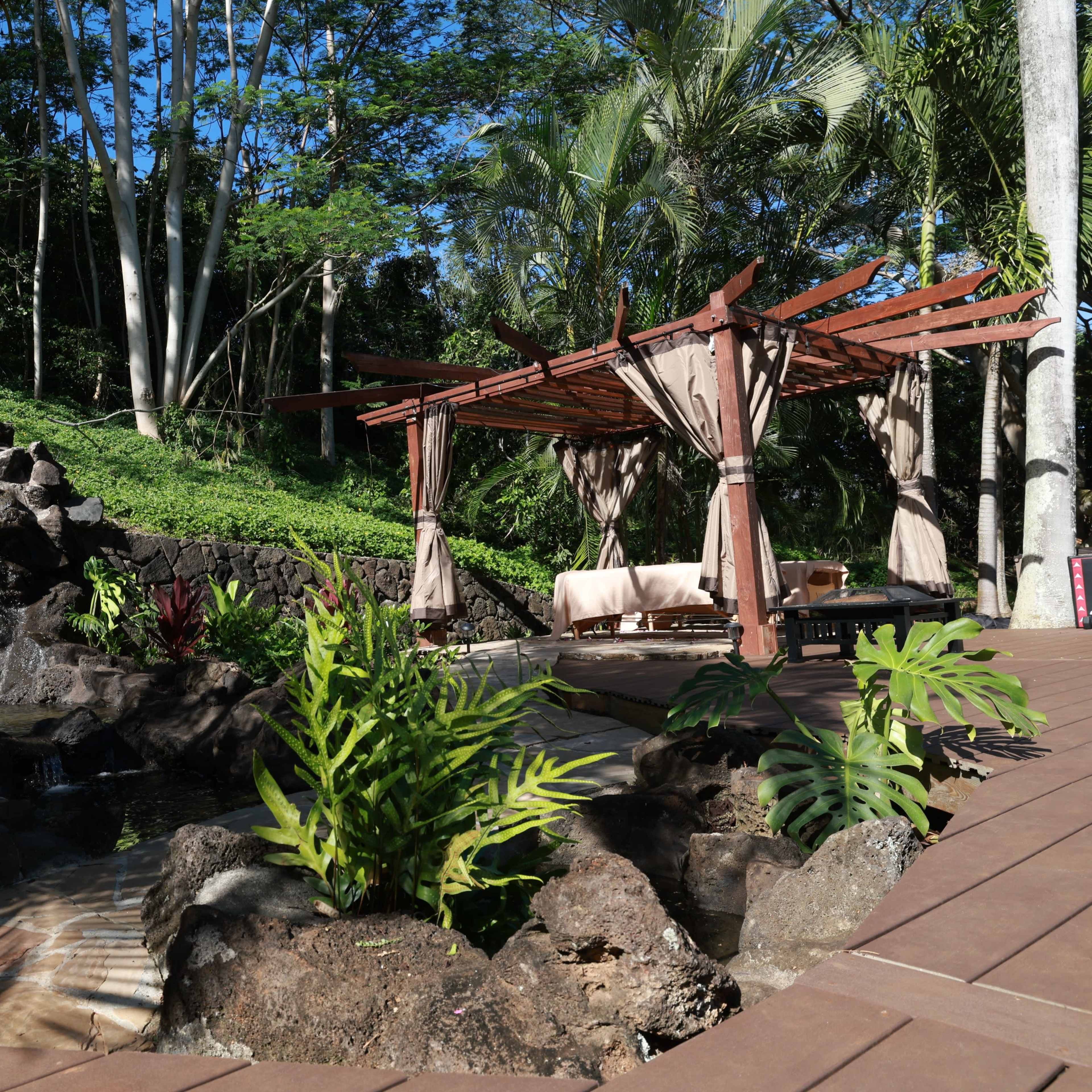 A wooden pergola with draped fabric overlooks a small waterfall surrounded by tropical plants and rock formations.