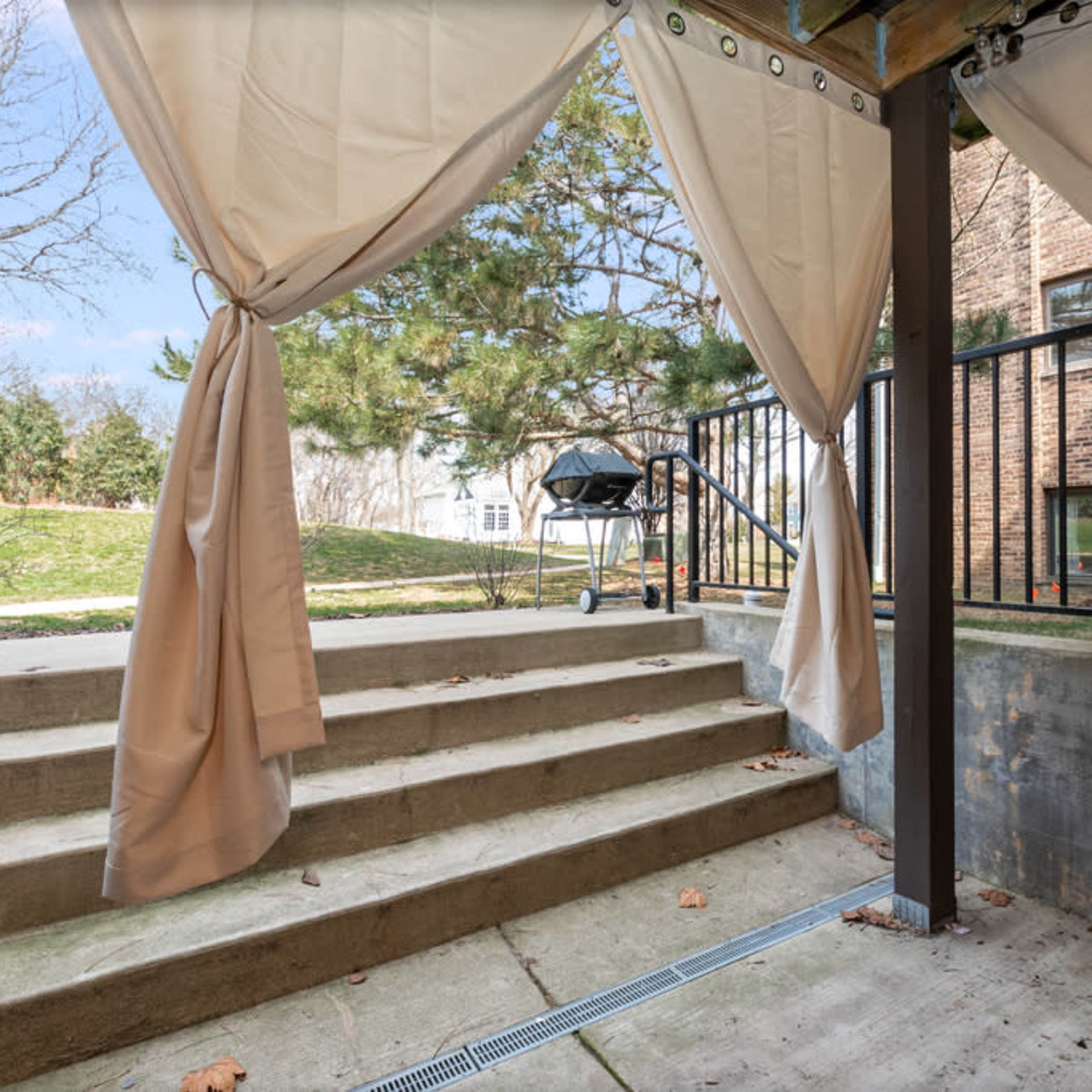A set of concrete steps leading up from a covered patio area adorned with draped curtains, with a barbecue grill positioned at the top of the steps.