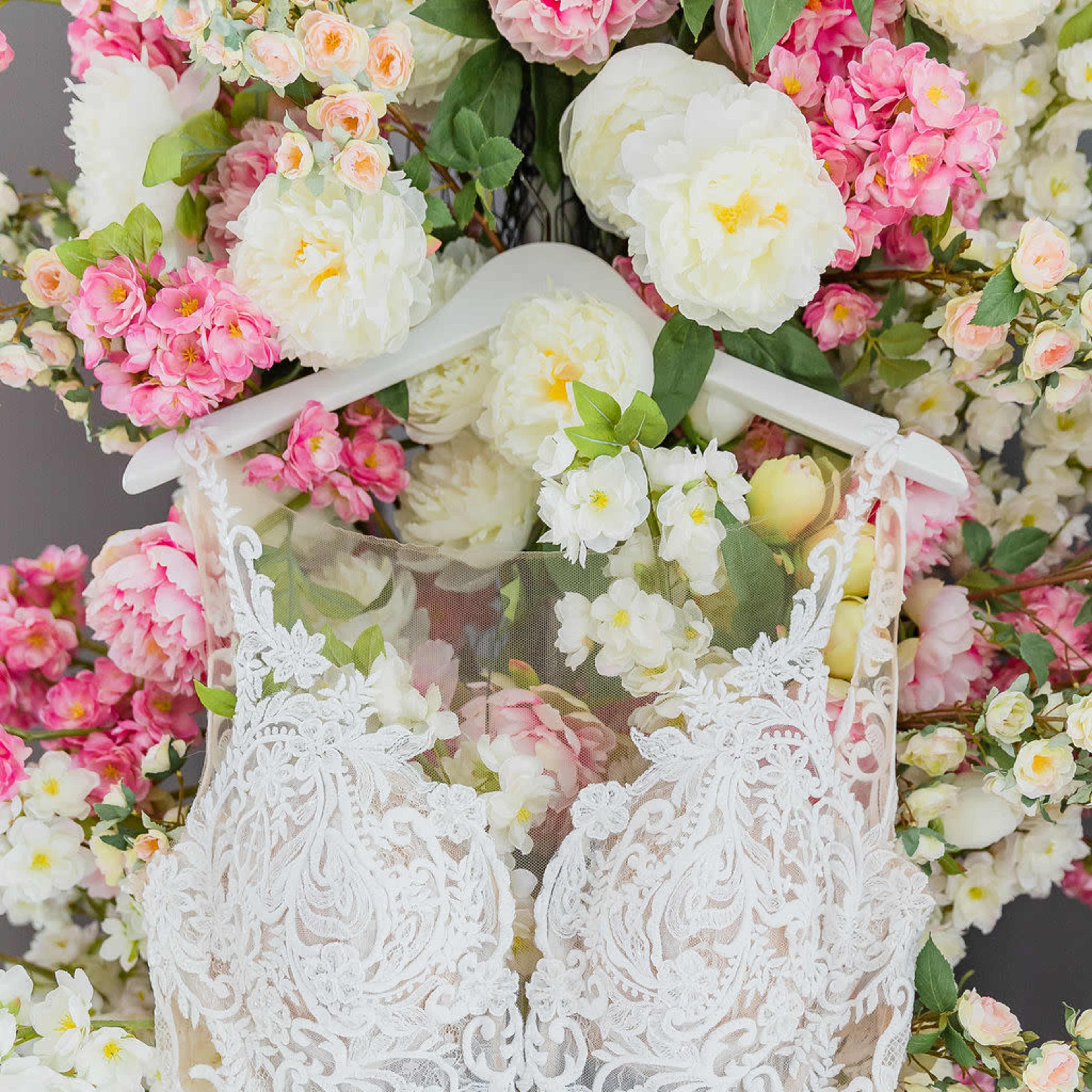 A white wedding dress with intricate lace detailing hangs against a backdrop of pink and white flowers.