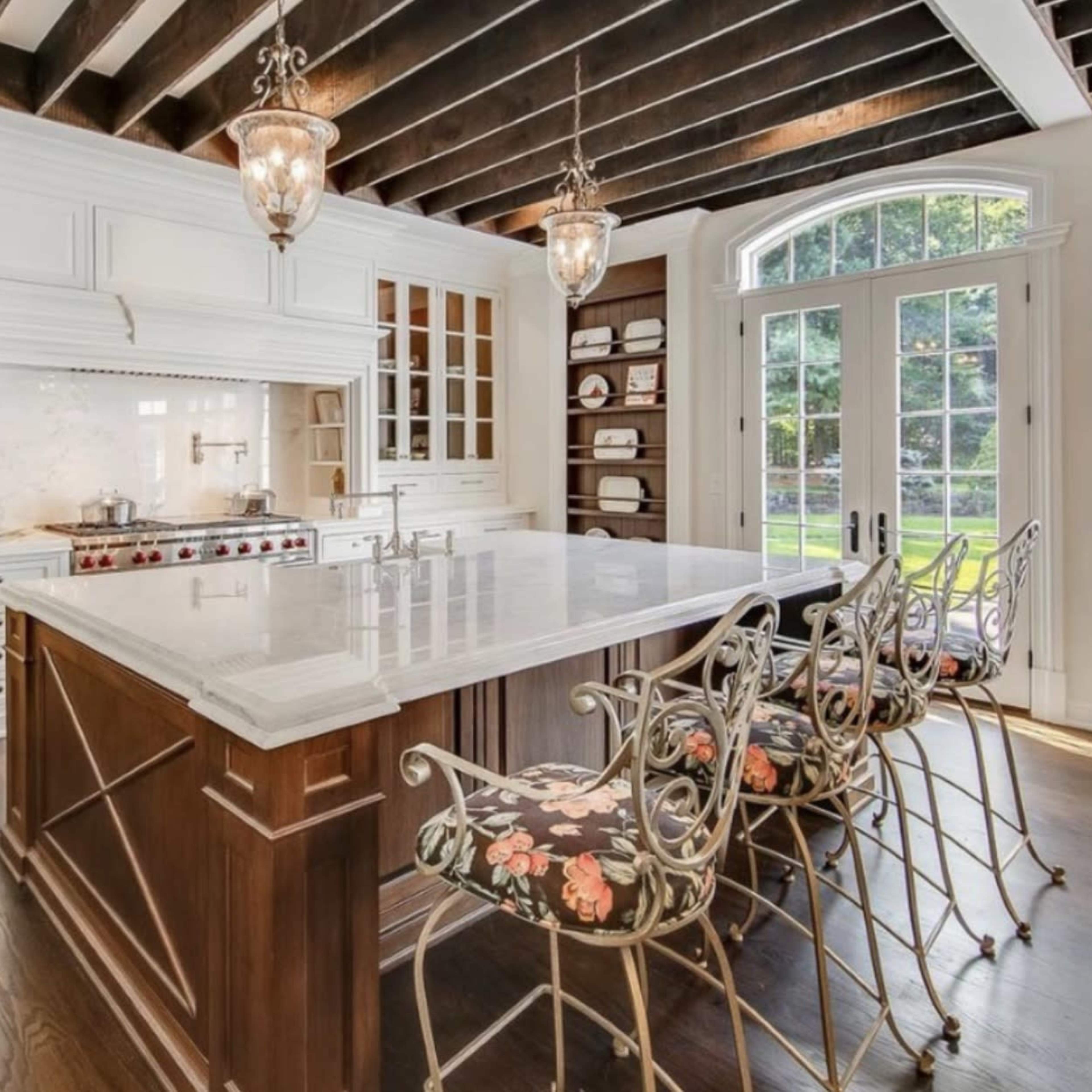 A modern kitchen with a large marble island, wooden beams on the ceiling, and glass doors leading to an outdoor area.
