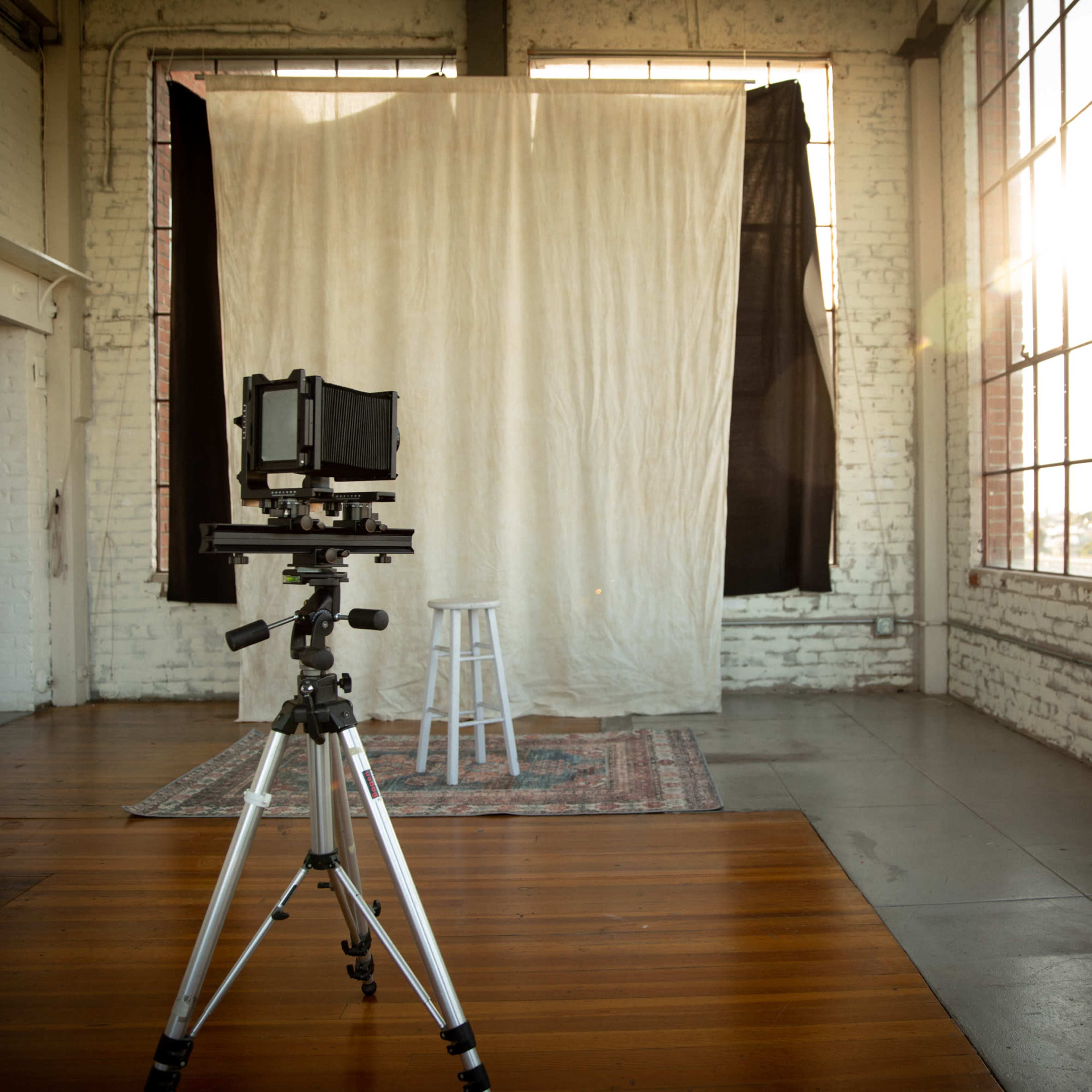 A photography studio featuring a large backdrop, a stool, and a camera mounted on a tripod, with sunlight streaming through large windows.