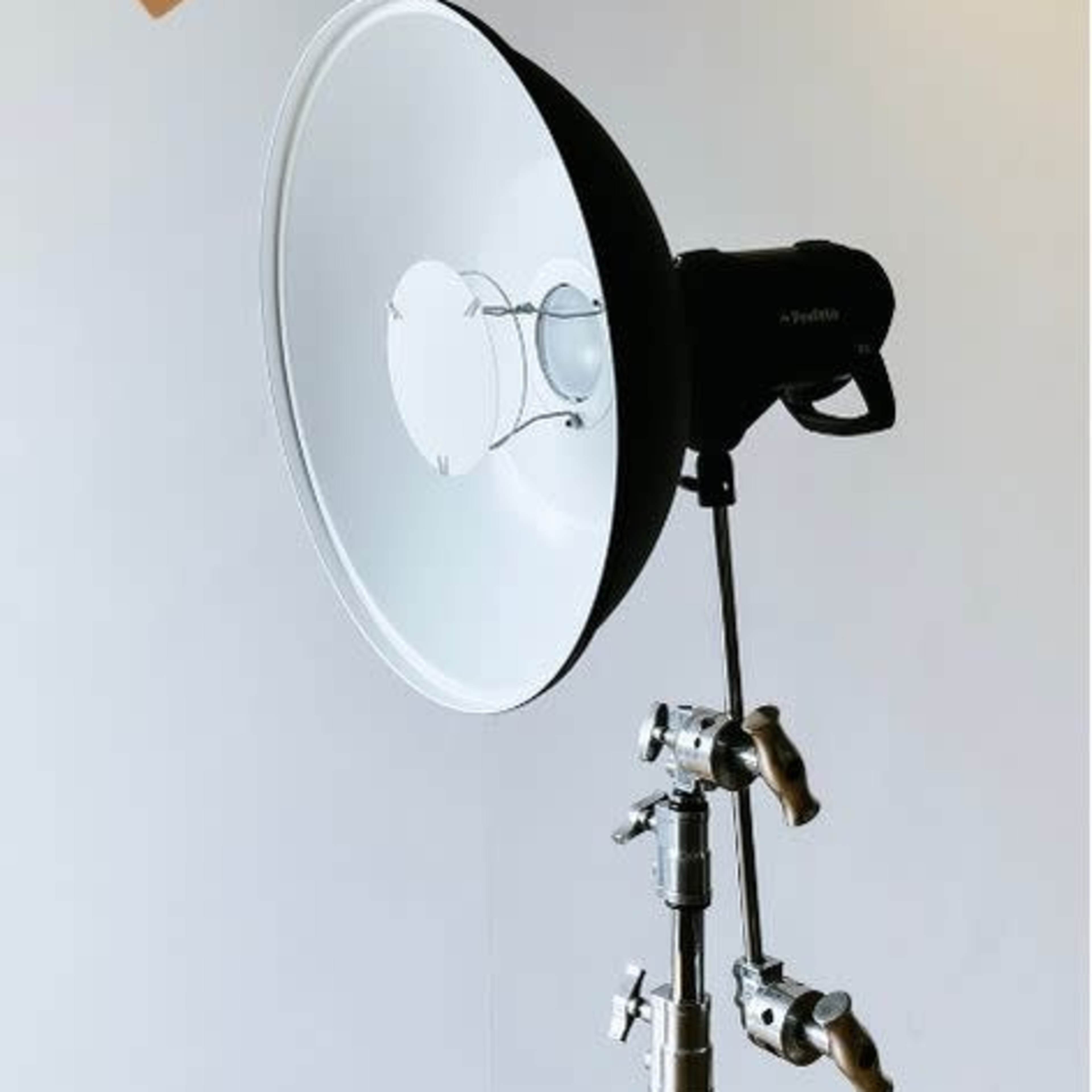 A studio light with a large reflector is mounted on a tripod stand against a plain wall.