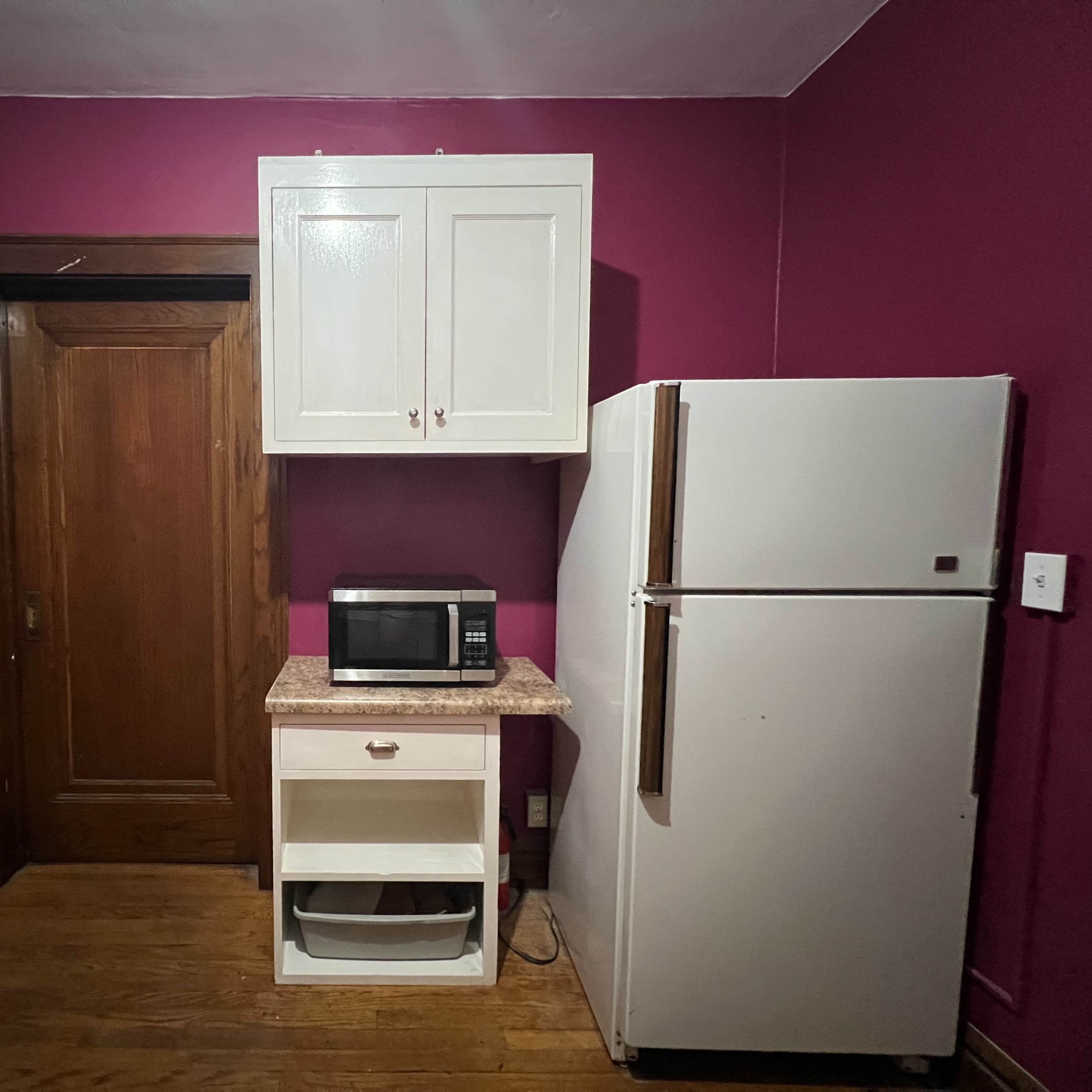 A kitchen corner with a purple wall, a wooden door, a white microwave on a countertop, and a white refrigerator.