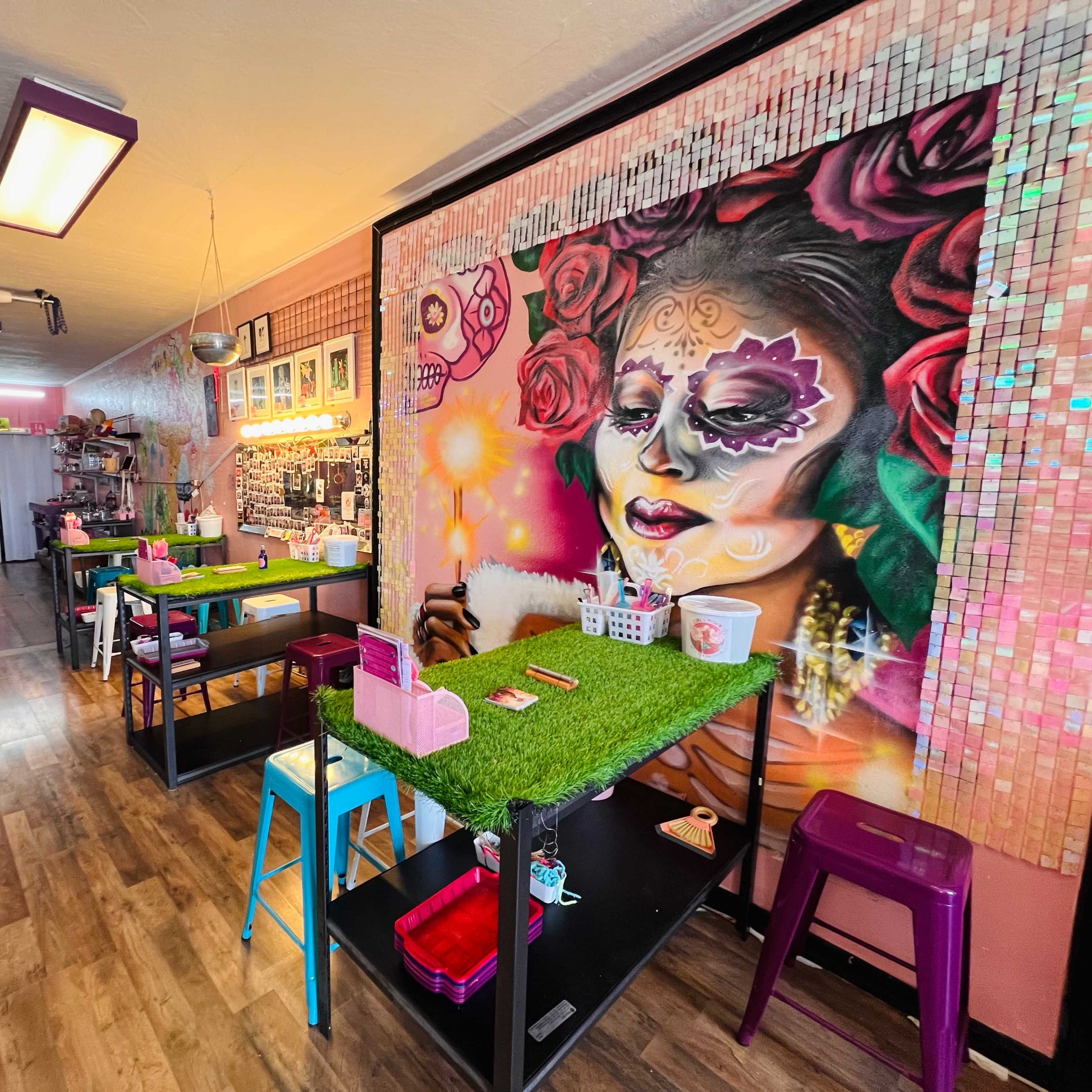 The interior of a colorful studio features tables with green turf tops, vibrant artwork on the wall, and shelves displaying various craft supplies.