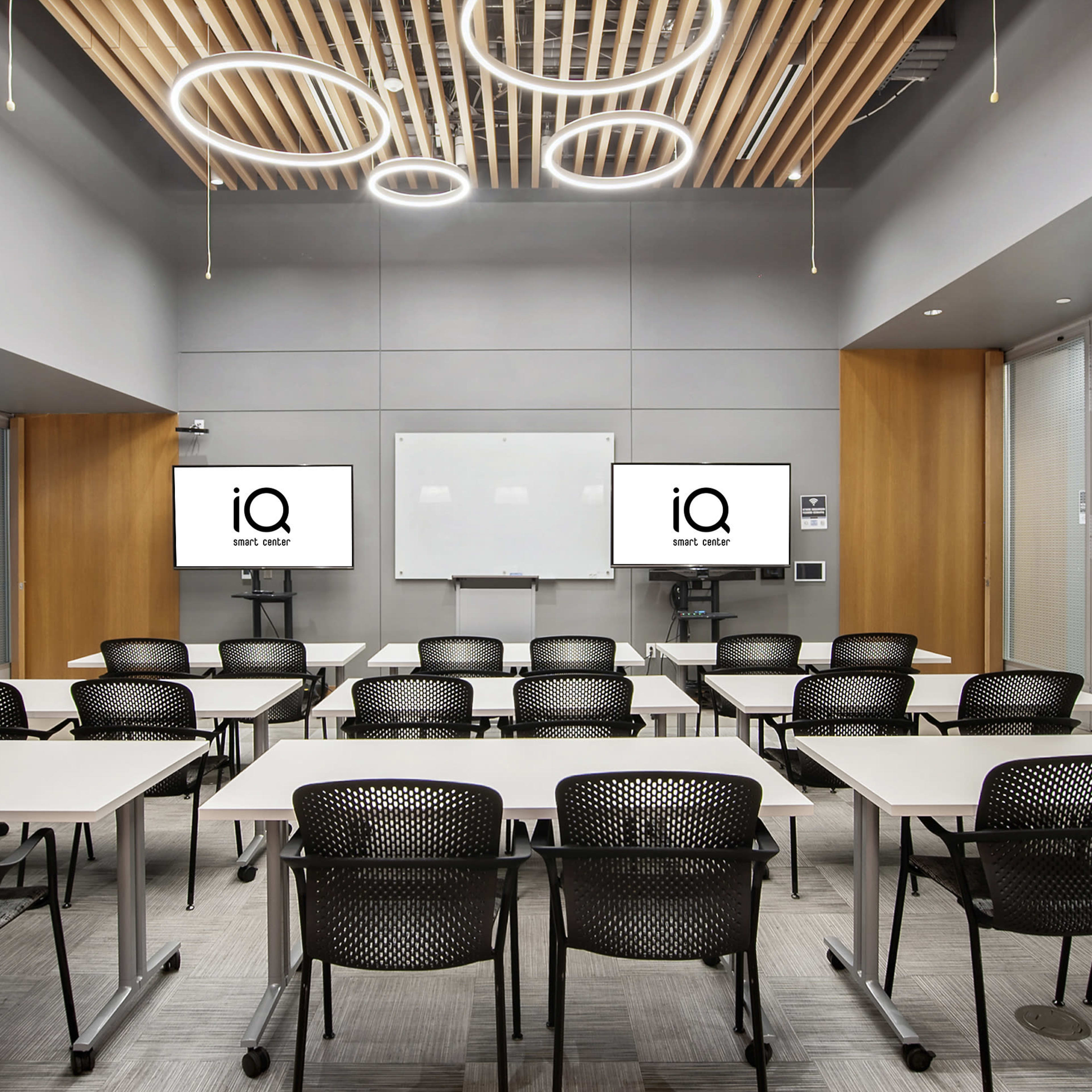 A modern classroom features white tables arranged in rows, black chairs, and two screens displaying a logo on the front wall.