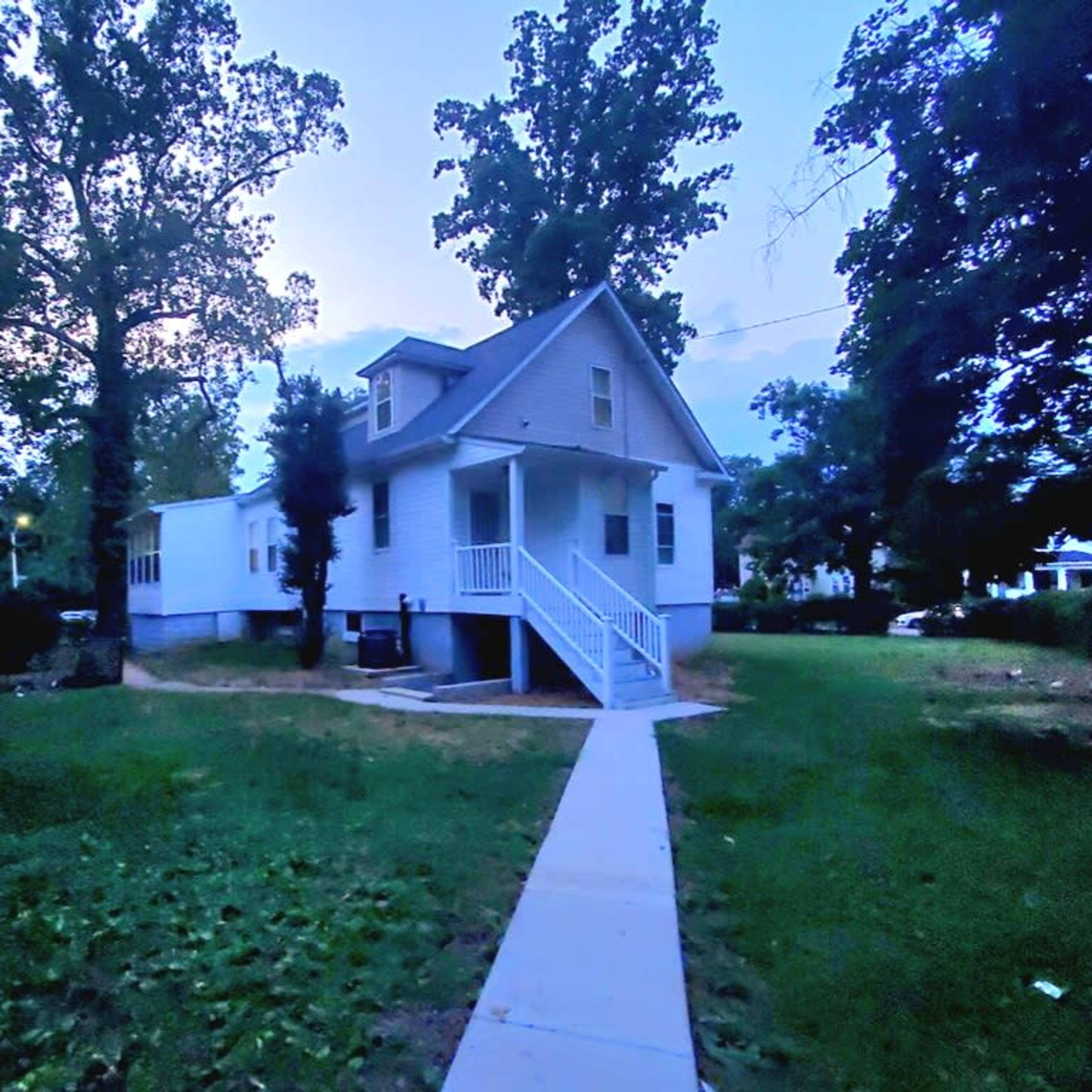 A two-story white house with a front porch is surrounded by a grassy yard and tall trees.