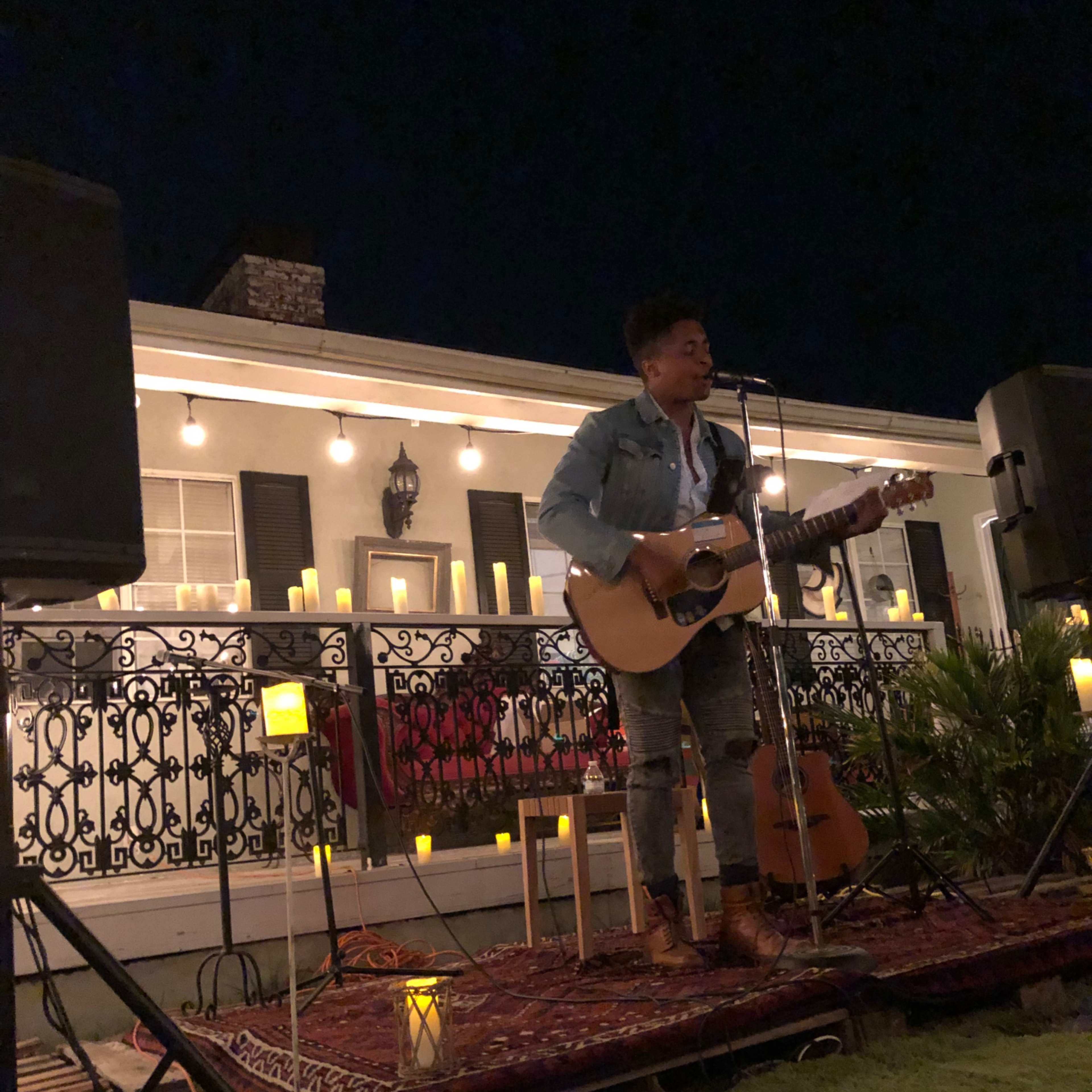 A musician performs on an outdoor stage with a guitar in front of an audience surrounded by candles and decorative lighting.