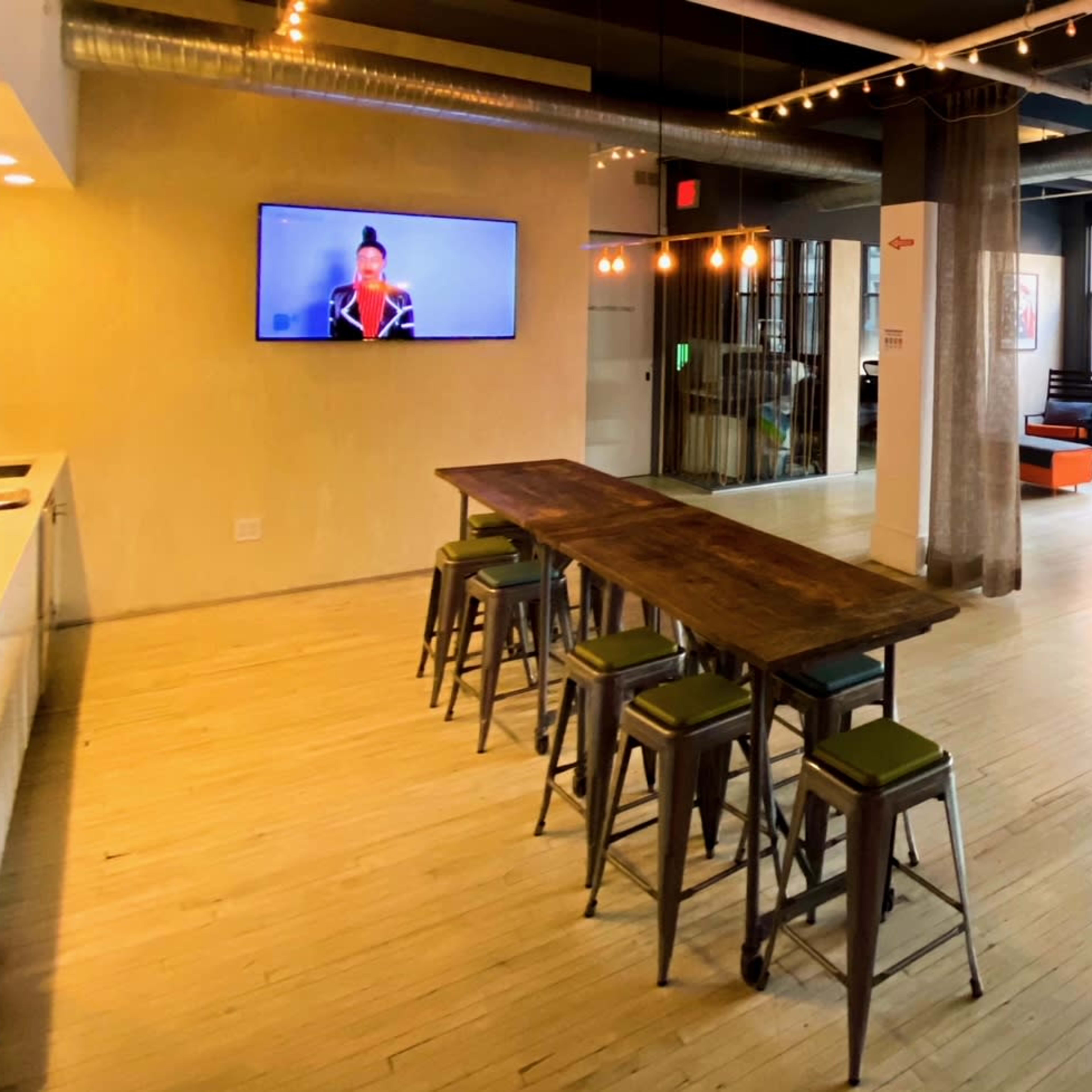 A spacious, modern open-plan living area with a long wooden dining table, bar stools, and a comfortable seating arrangement featuring a couch and armchairs, complemented by large windows and a television on the wall.