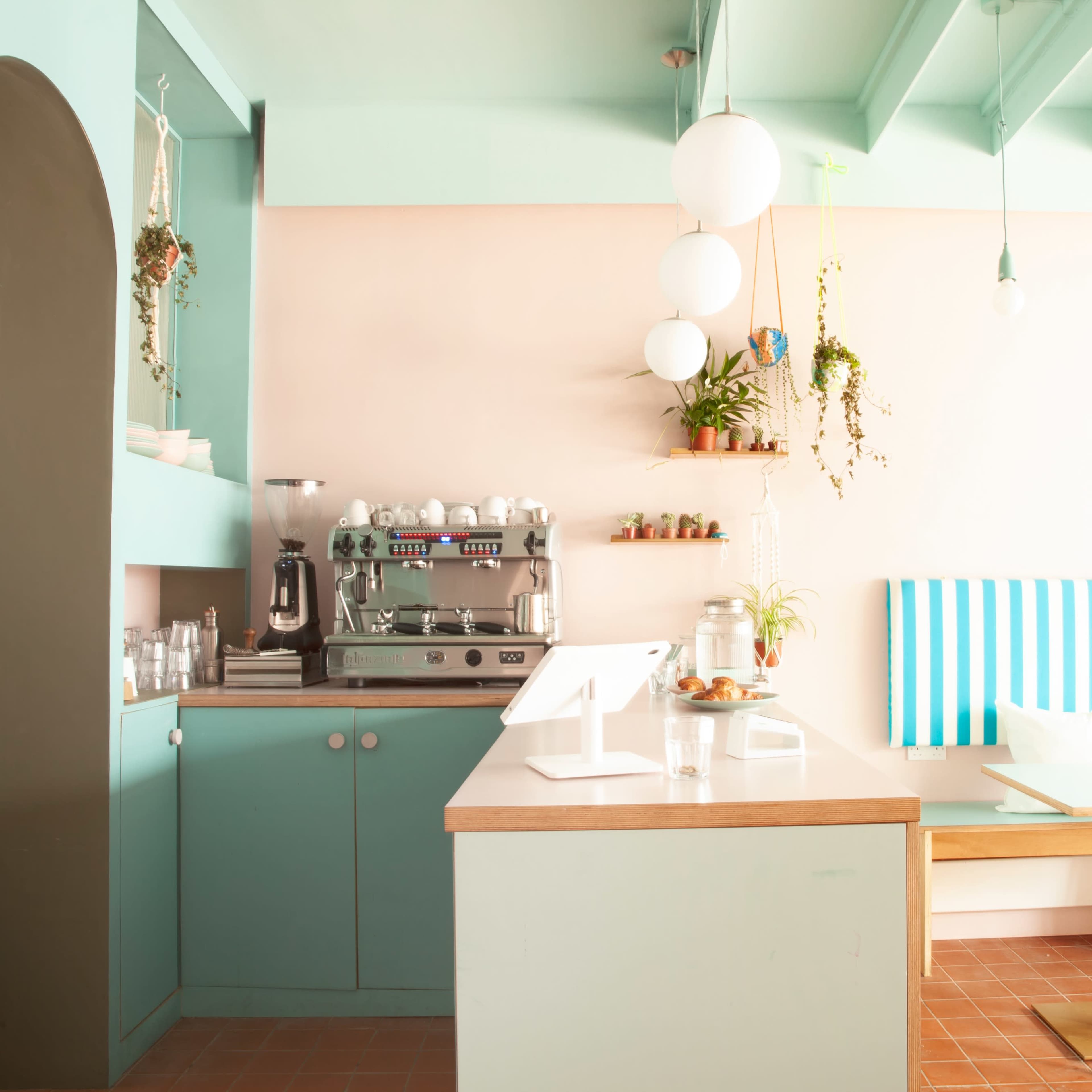 A brightly lit café features teal walls, a coffee machine, and wooden tables with striped seating.