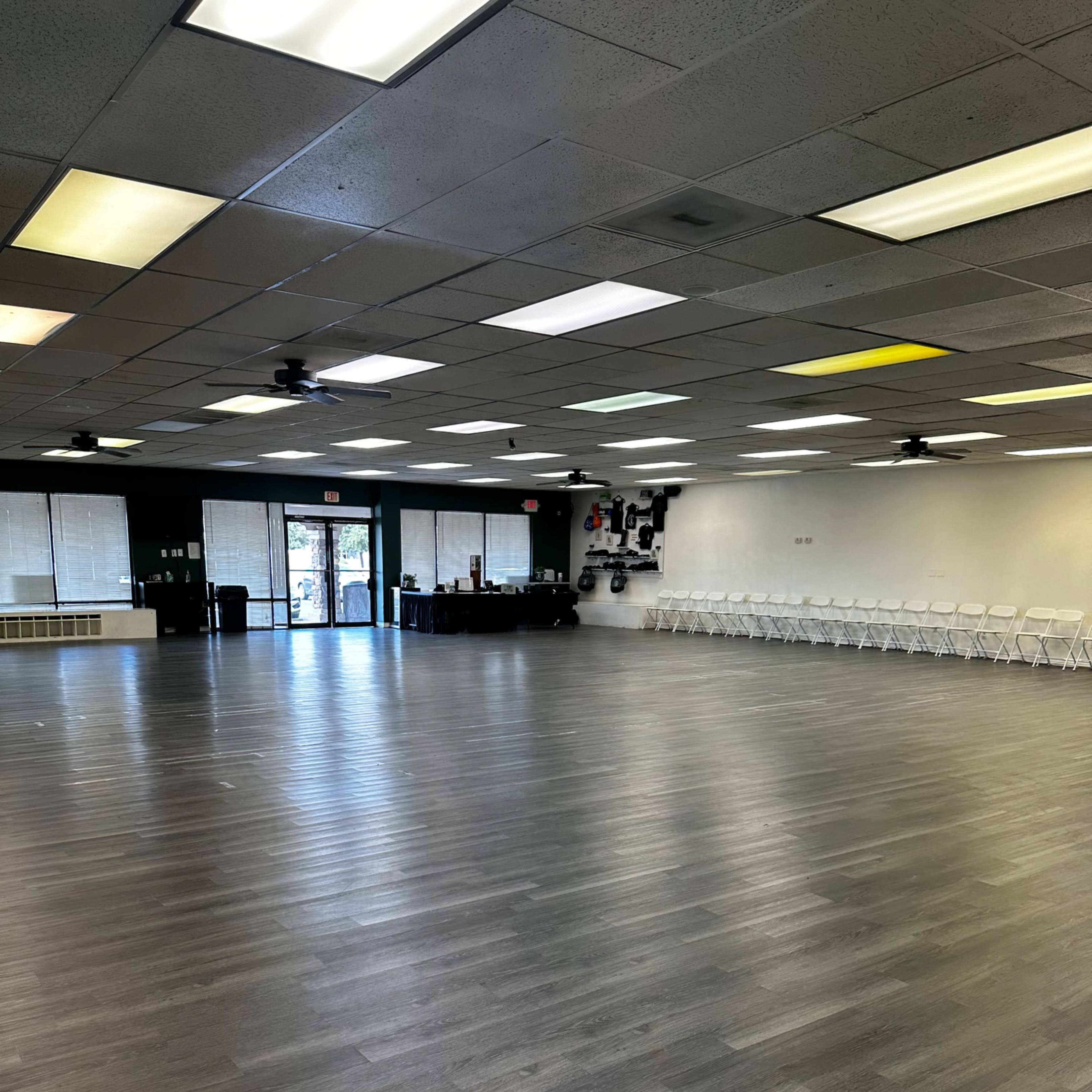 A spacious interior room with a wooden floor, empty chairs lined against one wall, and large windows providing natural light.