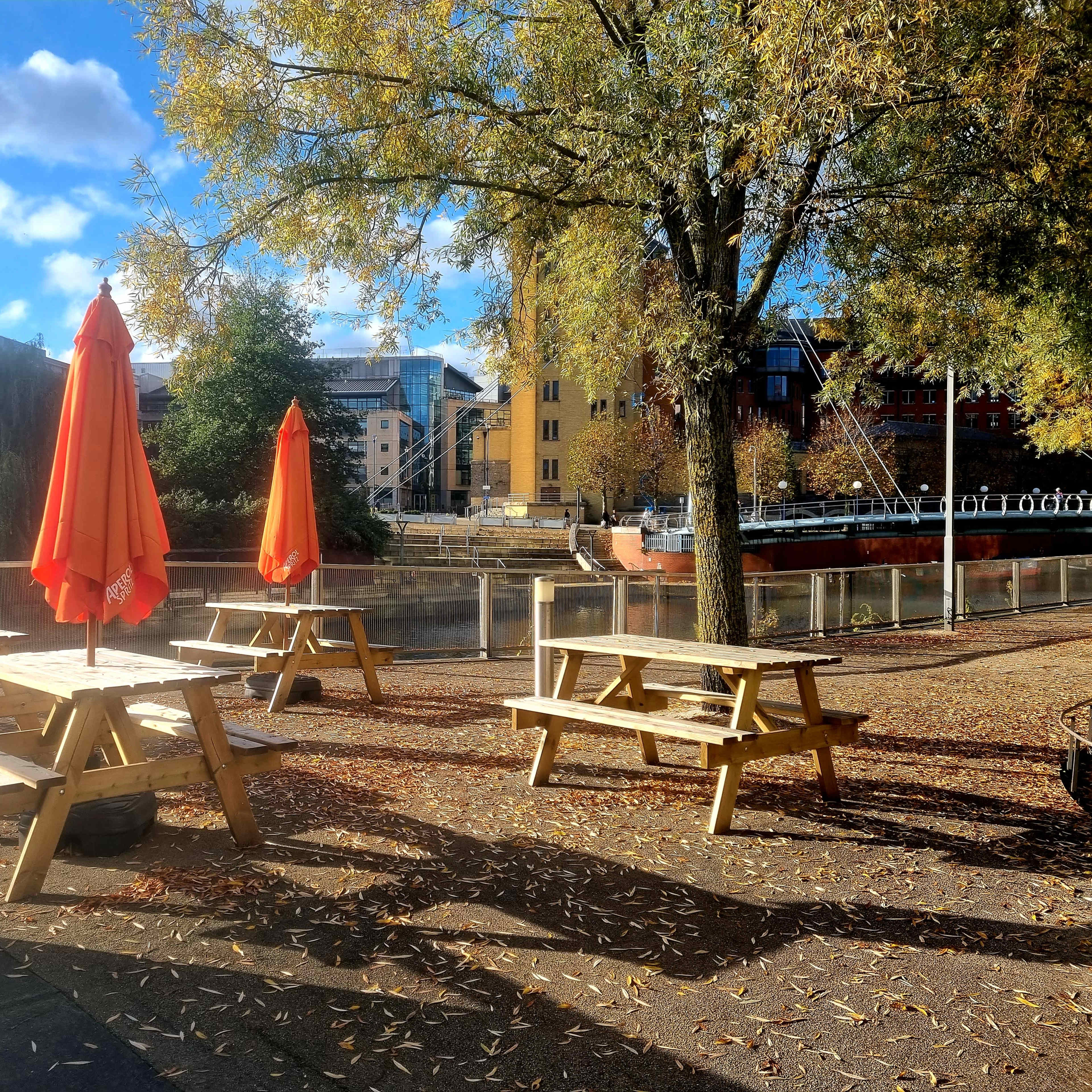 The scene features several wooden picnic tables under bright orange umbrellas near a calm river surrounded by trees and buildings.