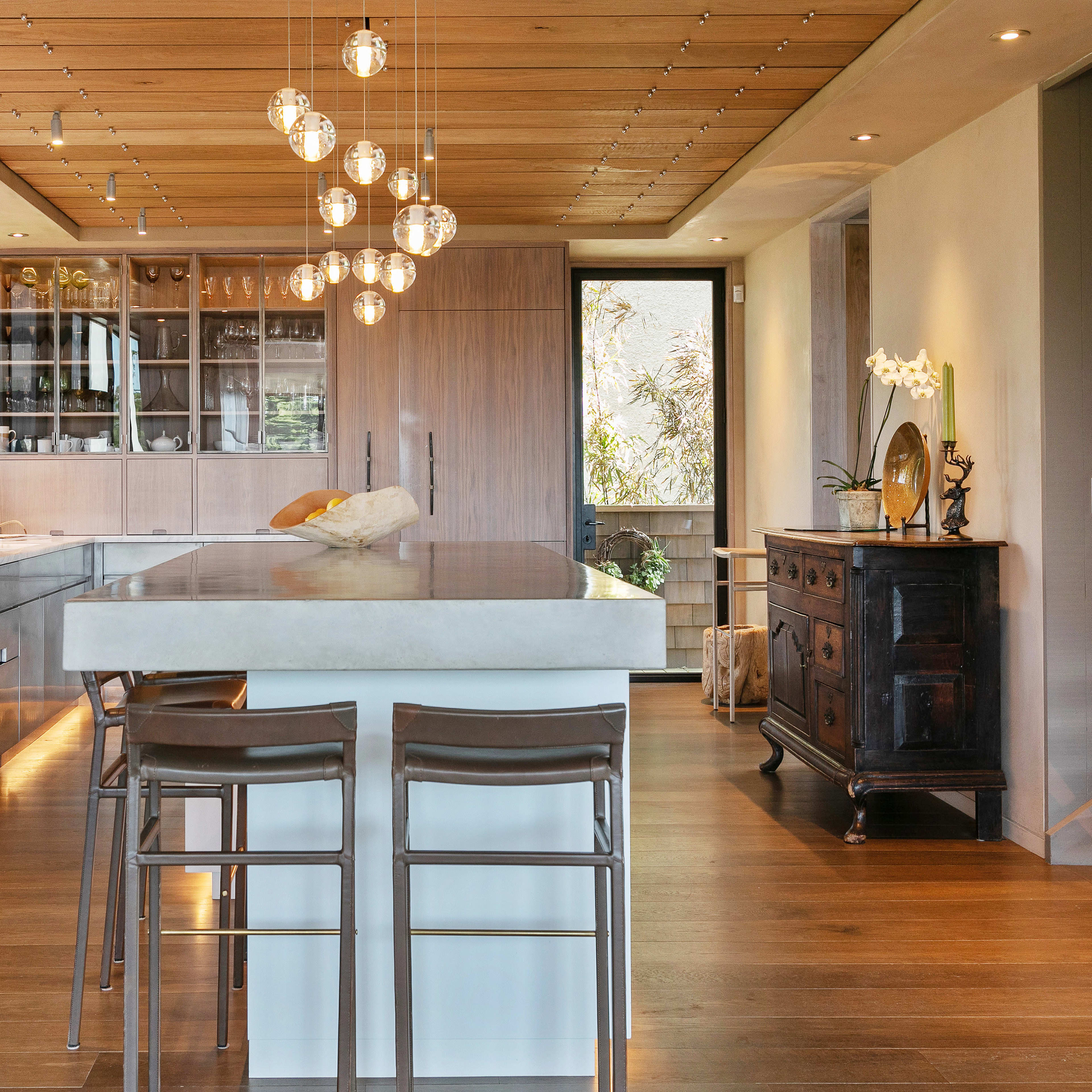 A modern kitchen features a long island with seating, pendant lighting, and large windows showing greenery outside.