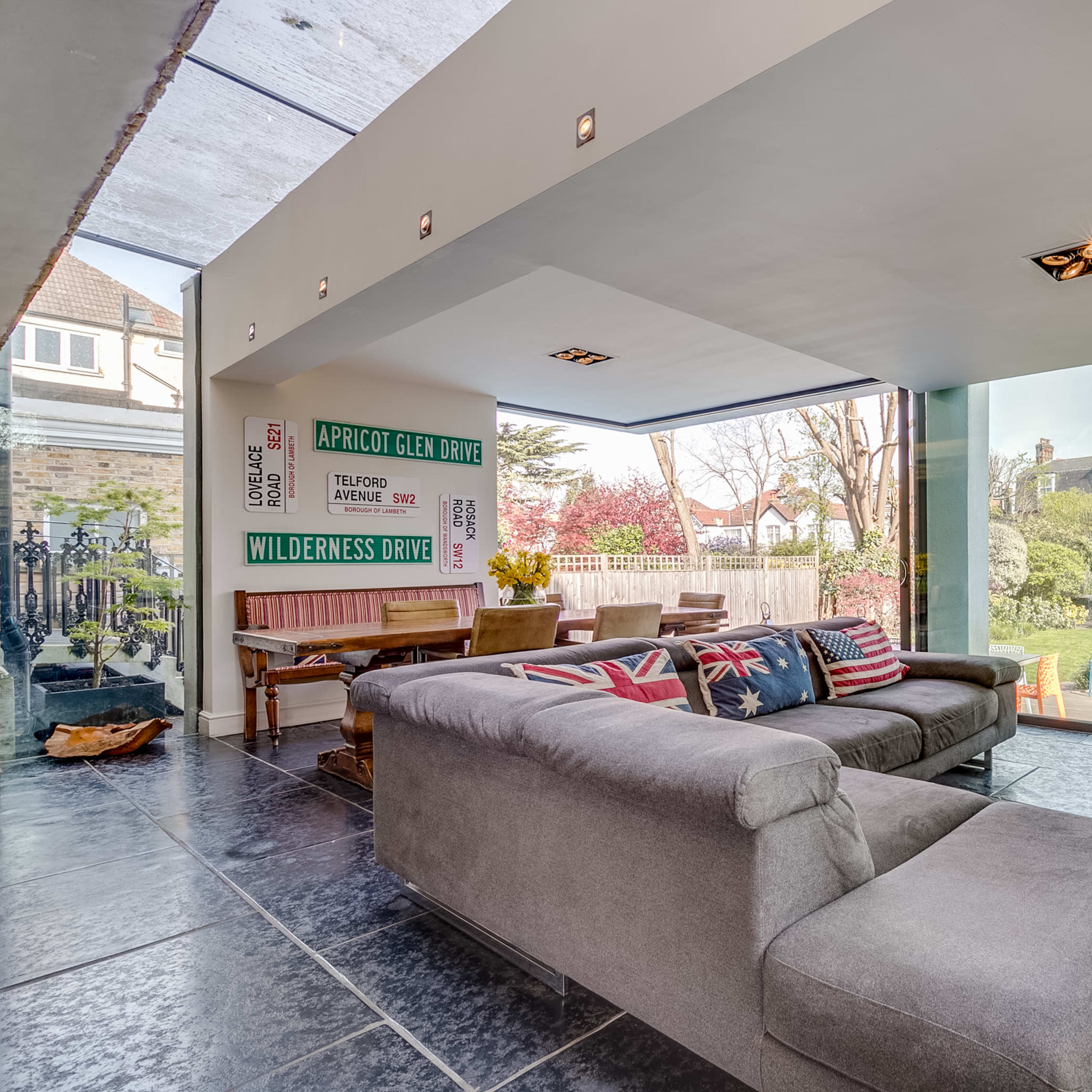 A modern living room with large glass windows overlooking a garden, featuring gray sofas and a wooden dining table.