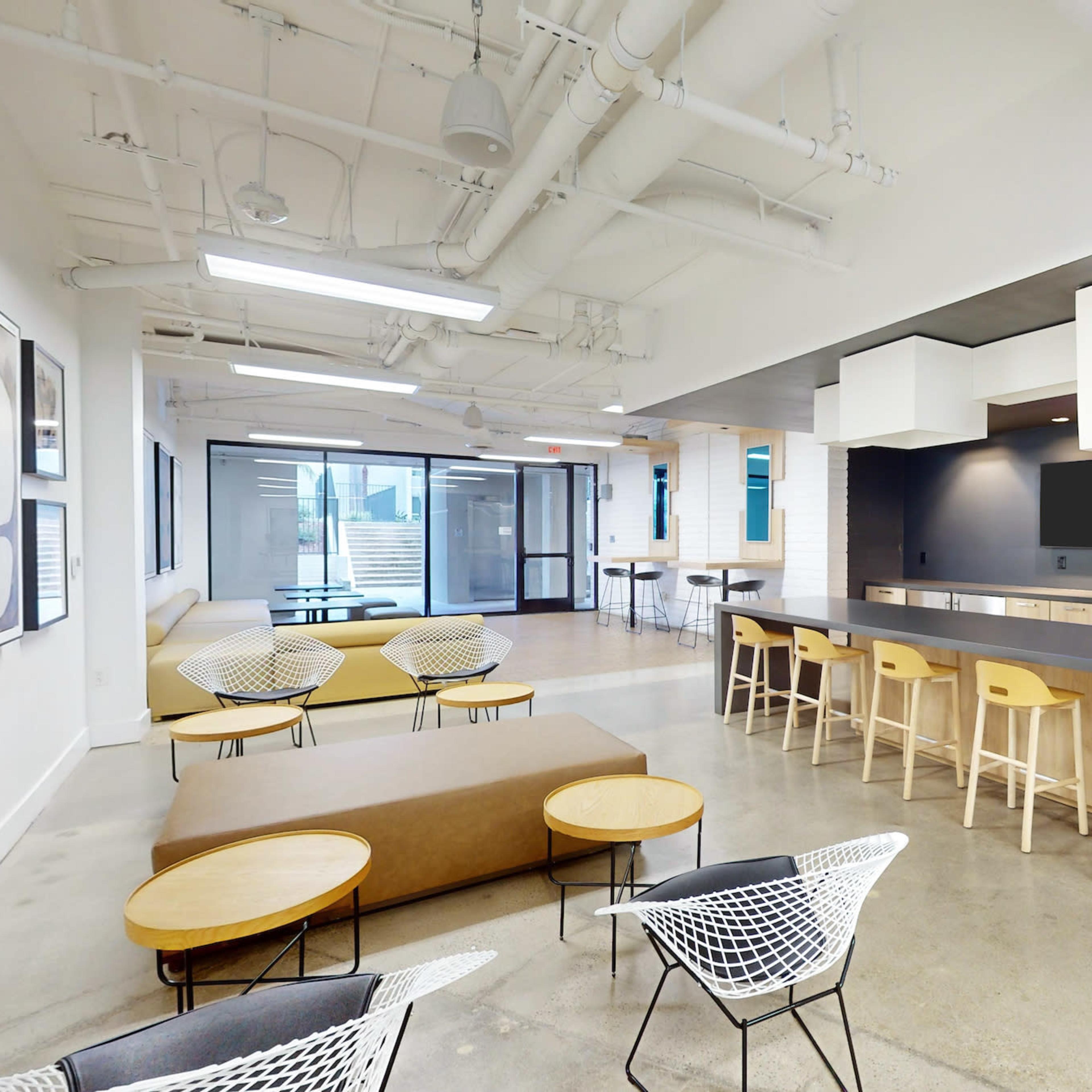 A modern office space featuring a kitchenette area with bar seating, several lounge chairs, and large windows leading to an outdoor area.