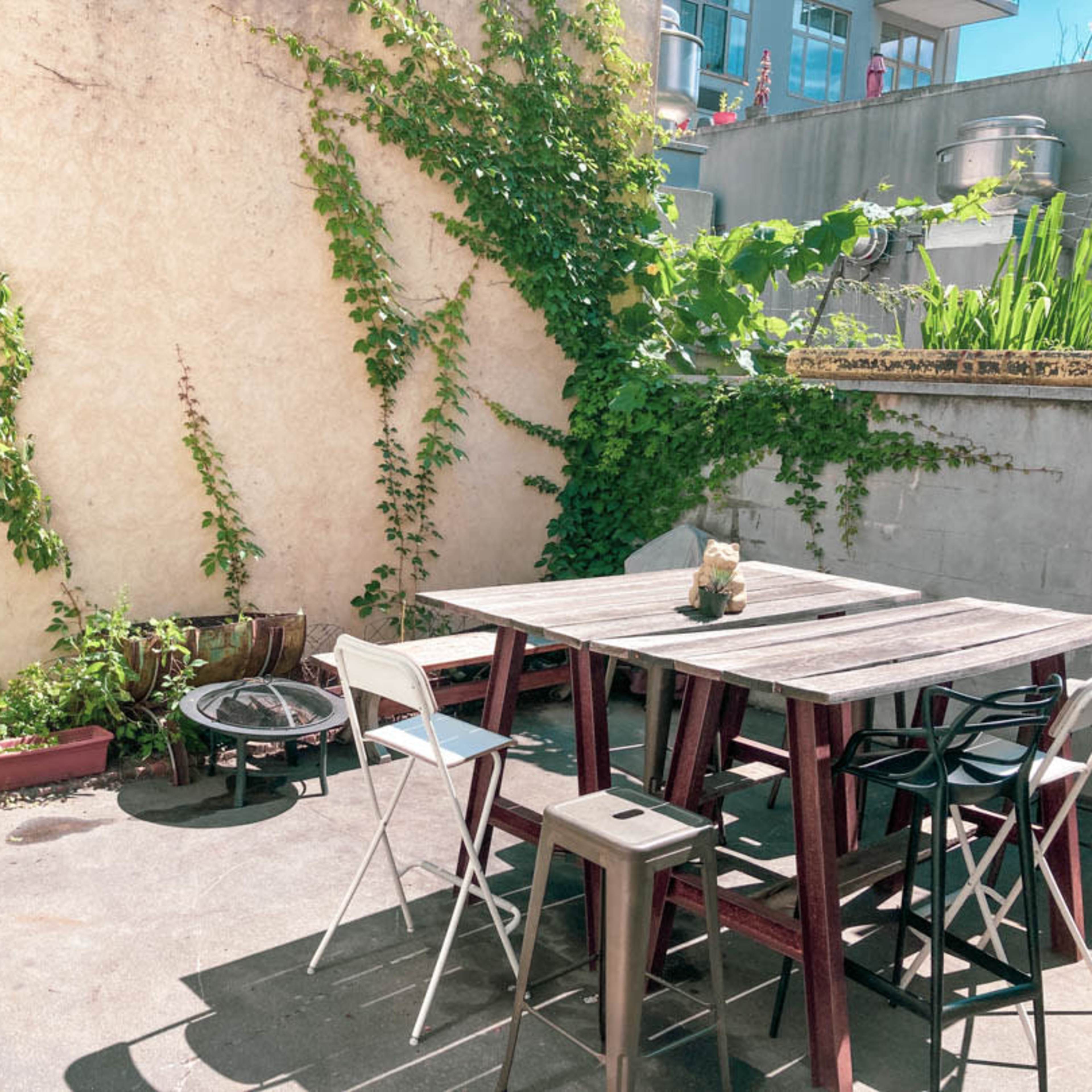 A small patio features a wooden table surrounded by metal chairs, with greenery climbing the wall and various plants in containers.