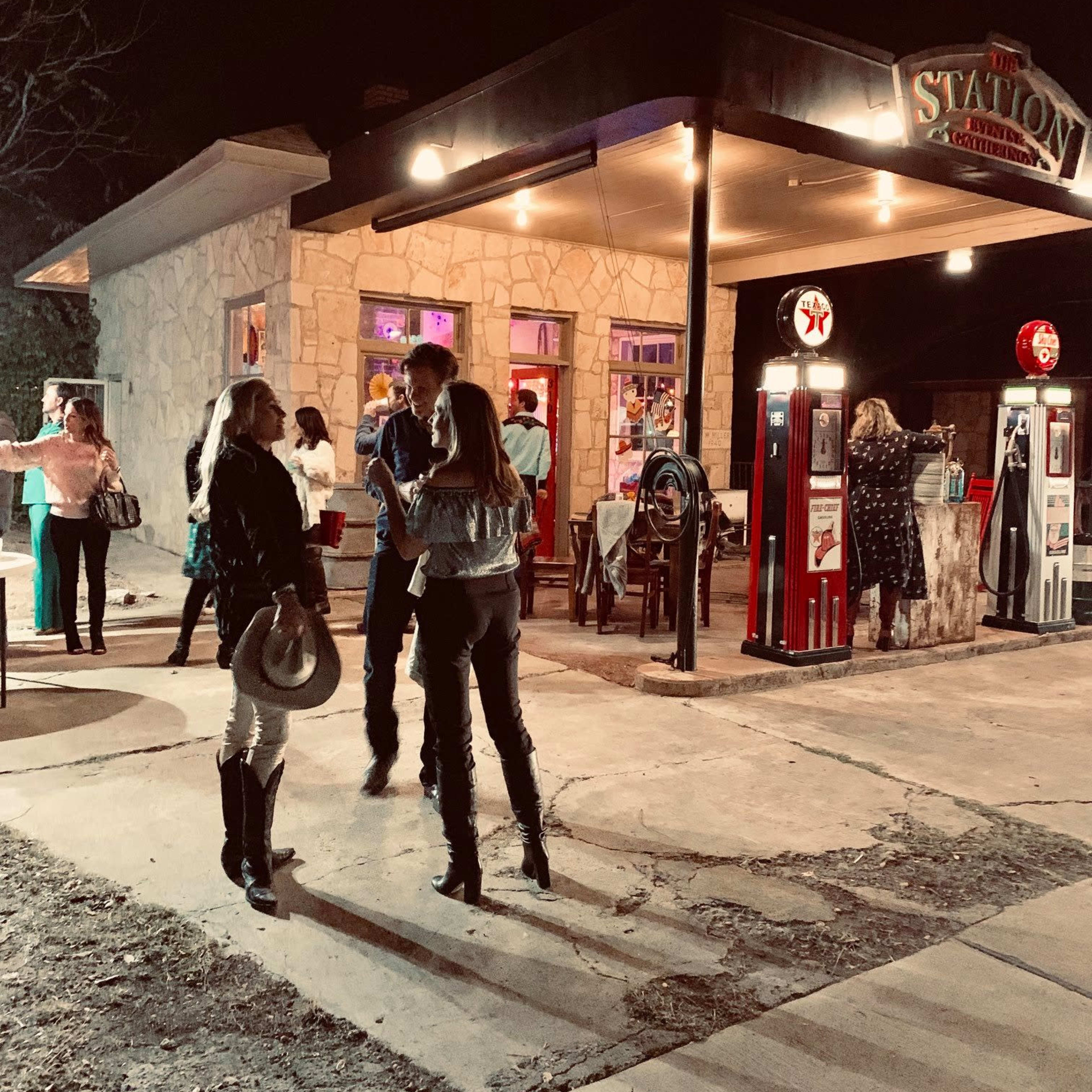 A group of people gather outside a vintage gas station at night, with colorful lights illuminating the stone building and antique gas pumps.