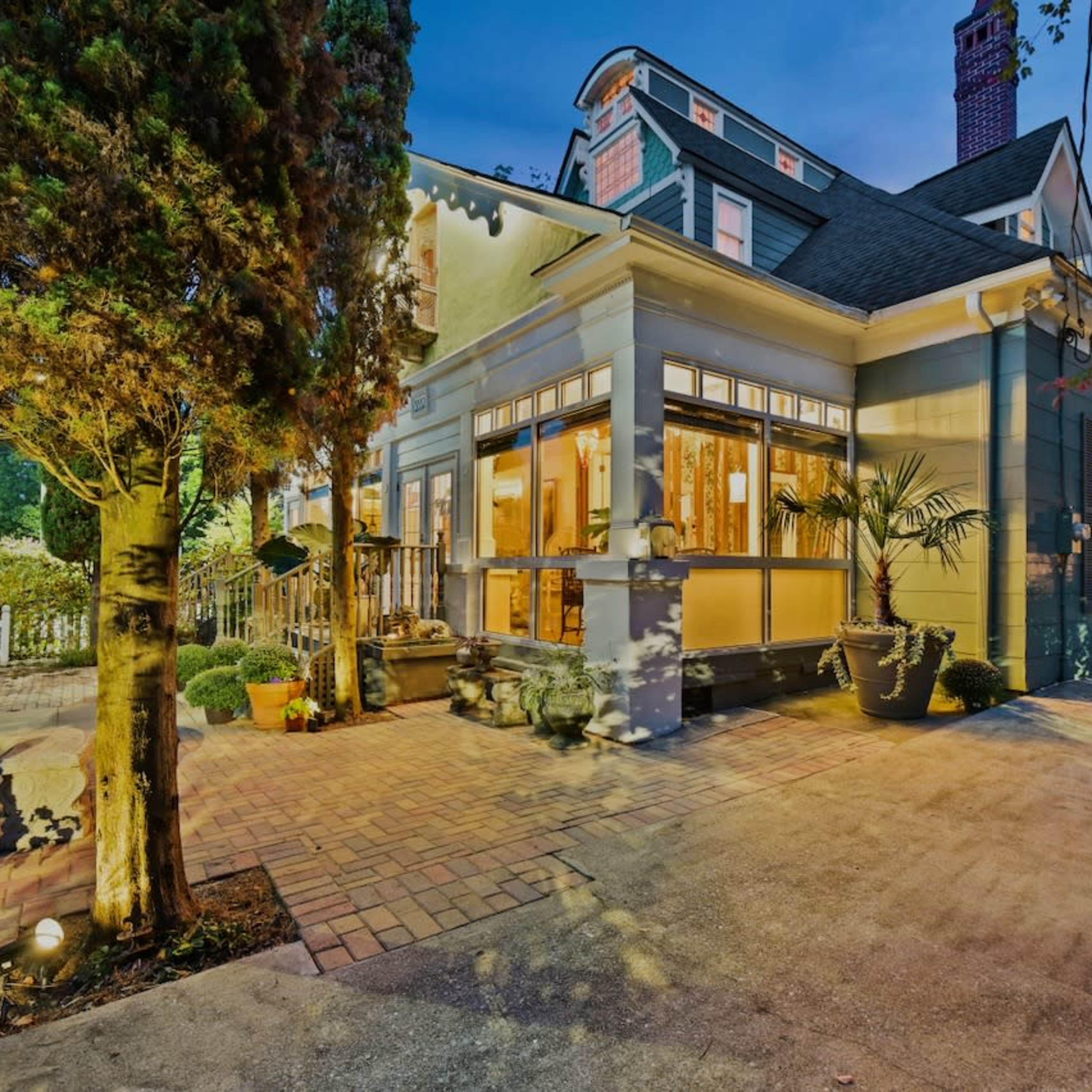A well-lit house with large windows and a landscaped entrance featuring potted plants and a brick pathway.