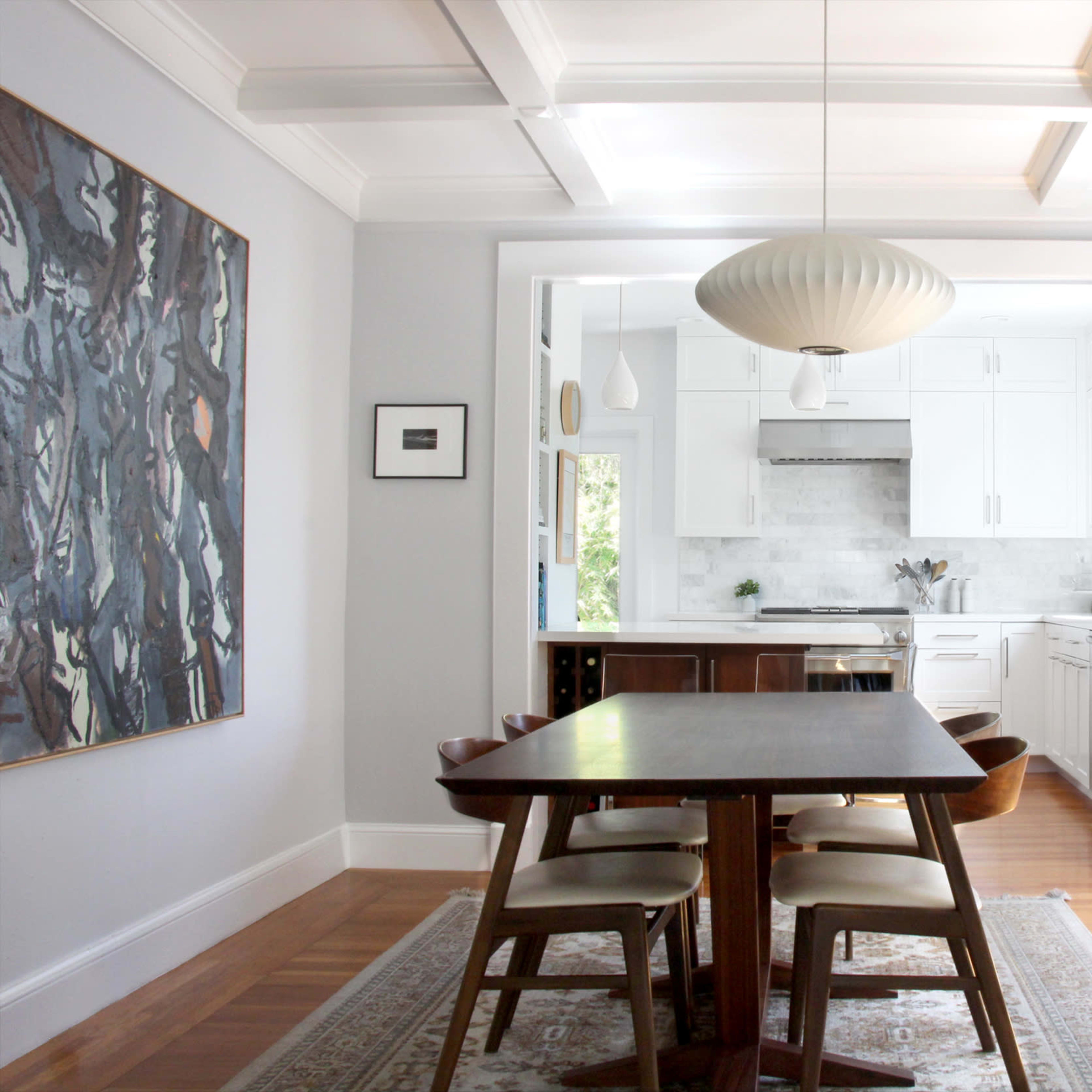 A dining area features a wooden table surrounded by chairs, with a modern light fixture hanging above and a view into a white kitchen through an open doorway.