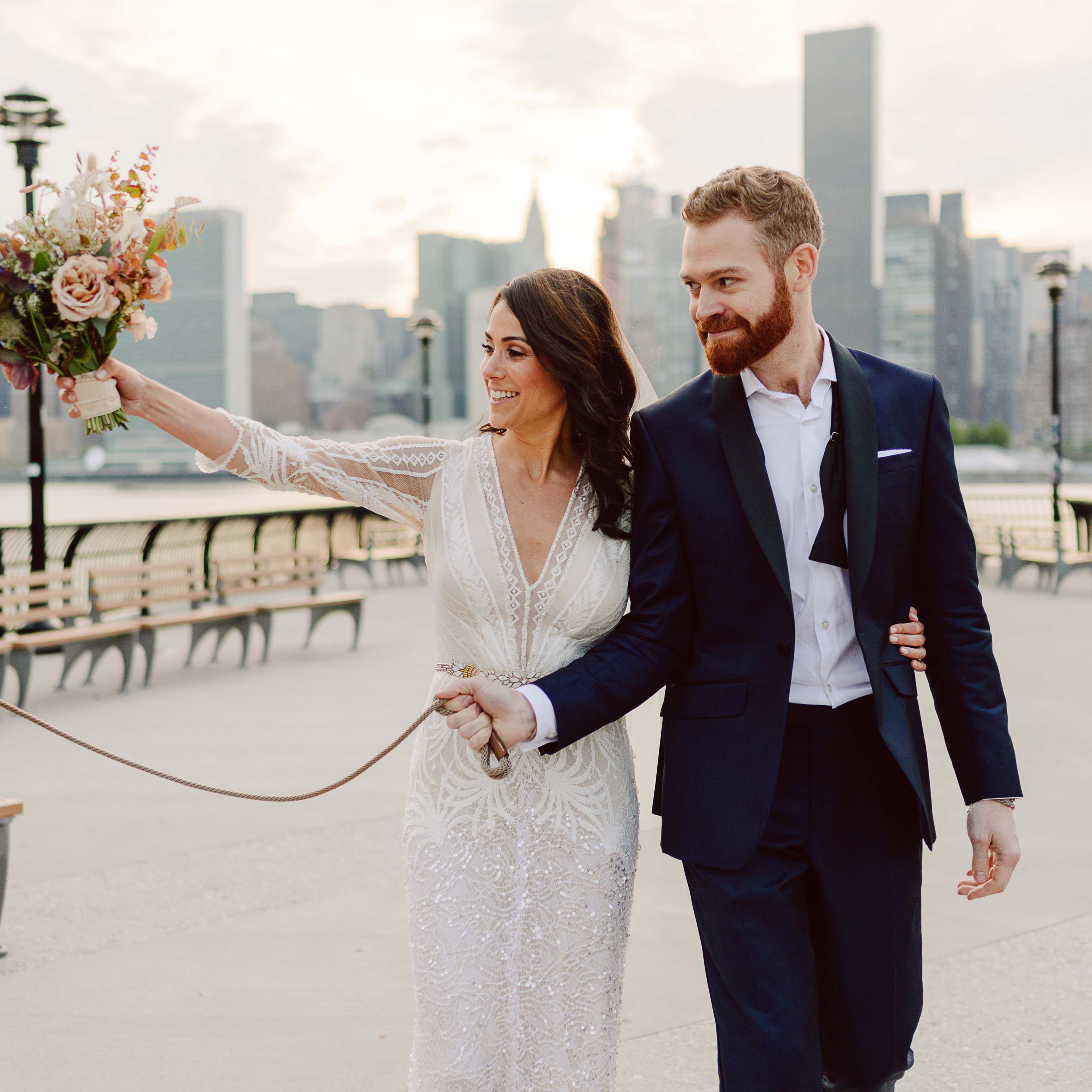 A couple dressed for a wedding leisurely walks together along a city waterfront, holding hands and a bouquet, with a dog jumping beside them.