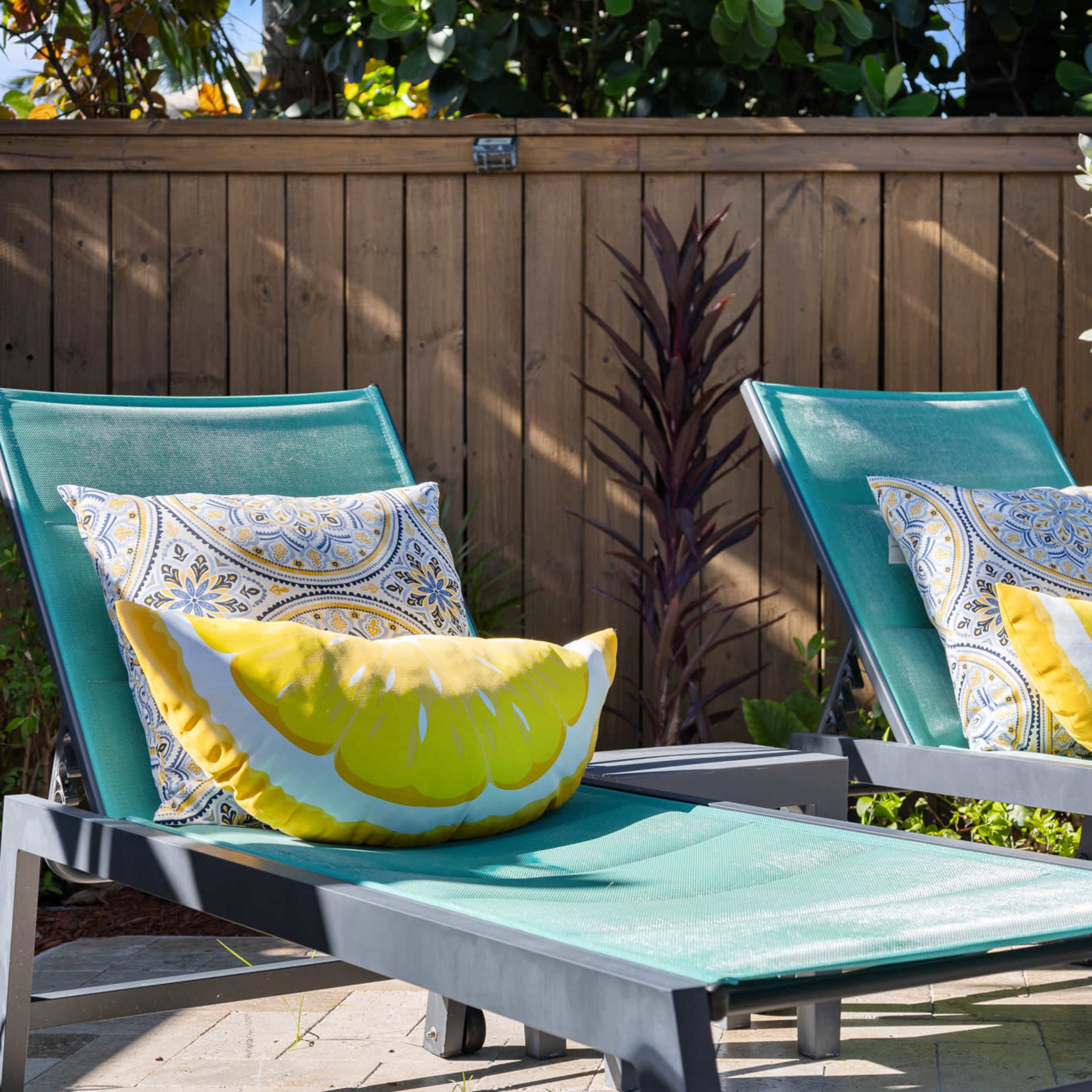 Two green lounge chairs with decorative yellow and patterned pillows are positioned next to a wooden fenced area filled with plants.