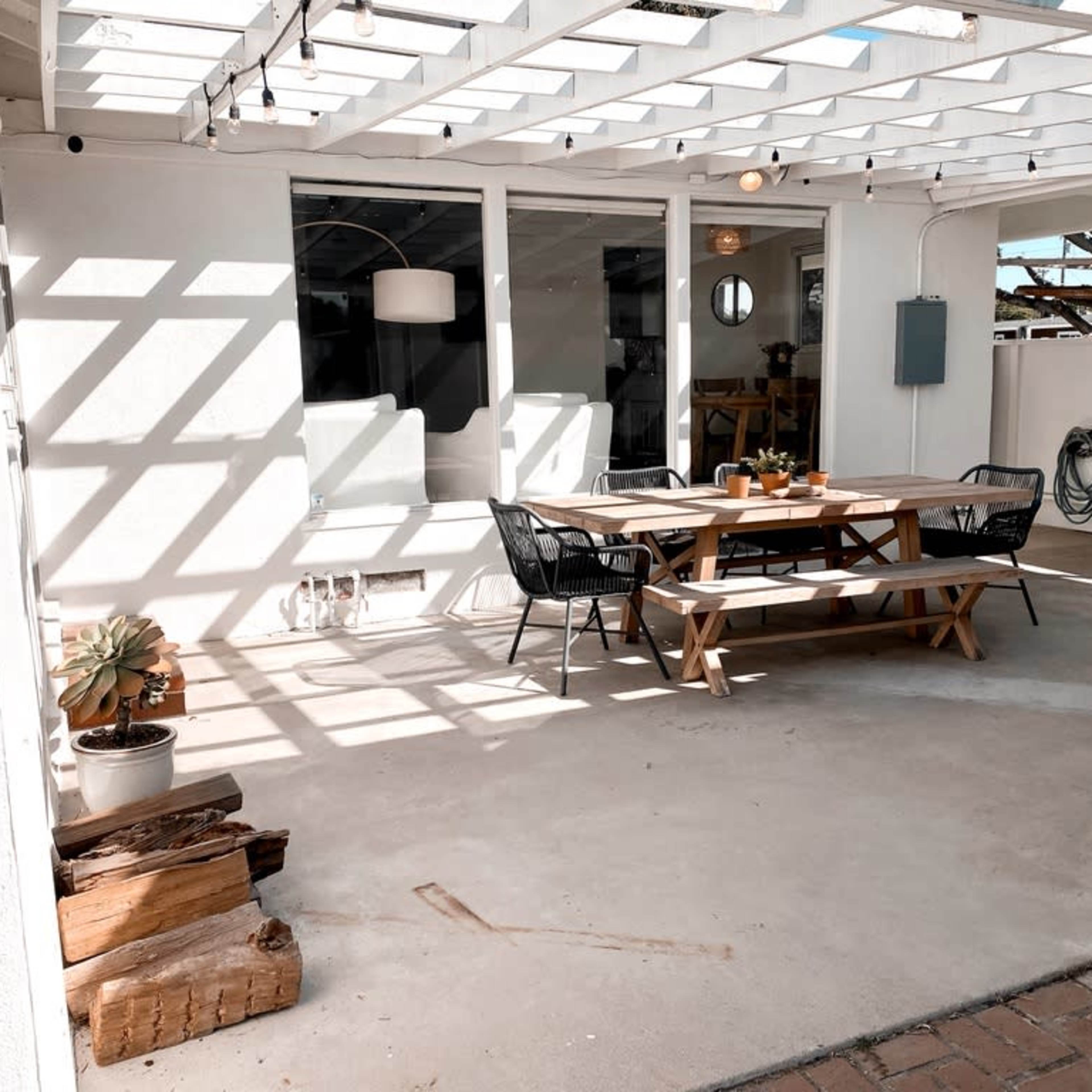 A patio area features a wooden table with benches, surrounded by potted plants and sunlight casting shadows through a slatted overhead structure.
