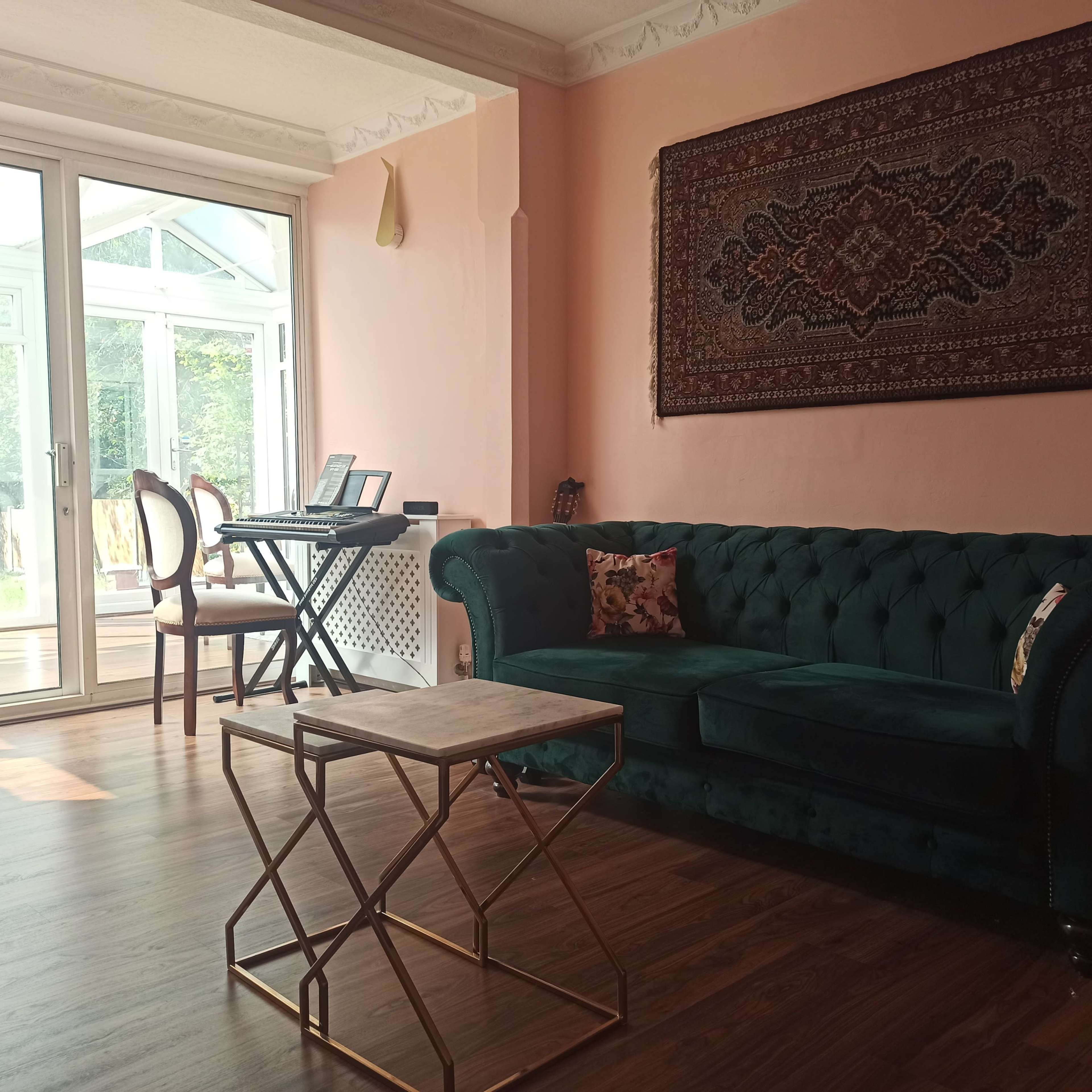 A spacious room with a green tufted sofa, a small gold coffee table, and a glass door leading to a sunlit conservatory.