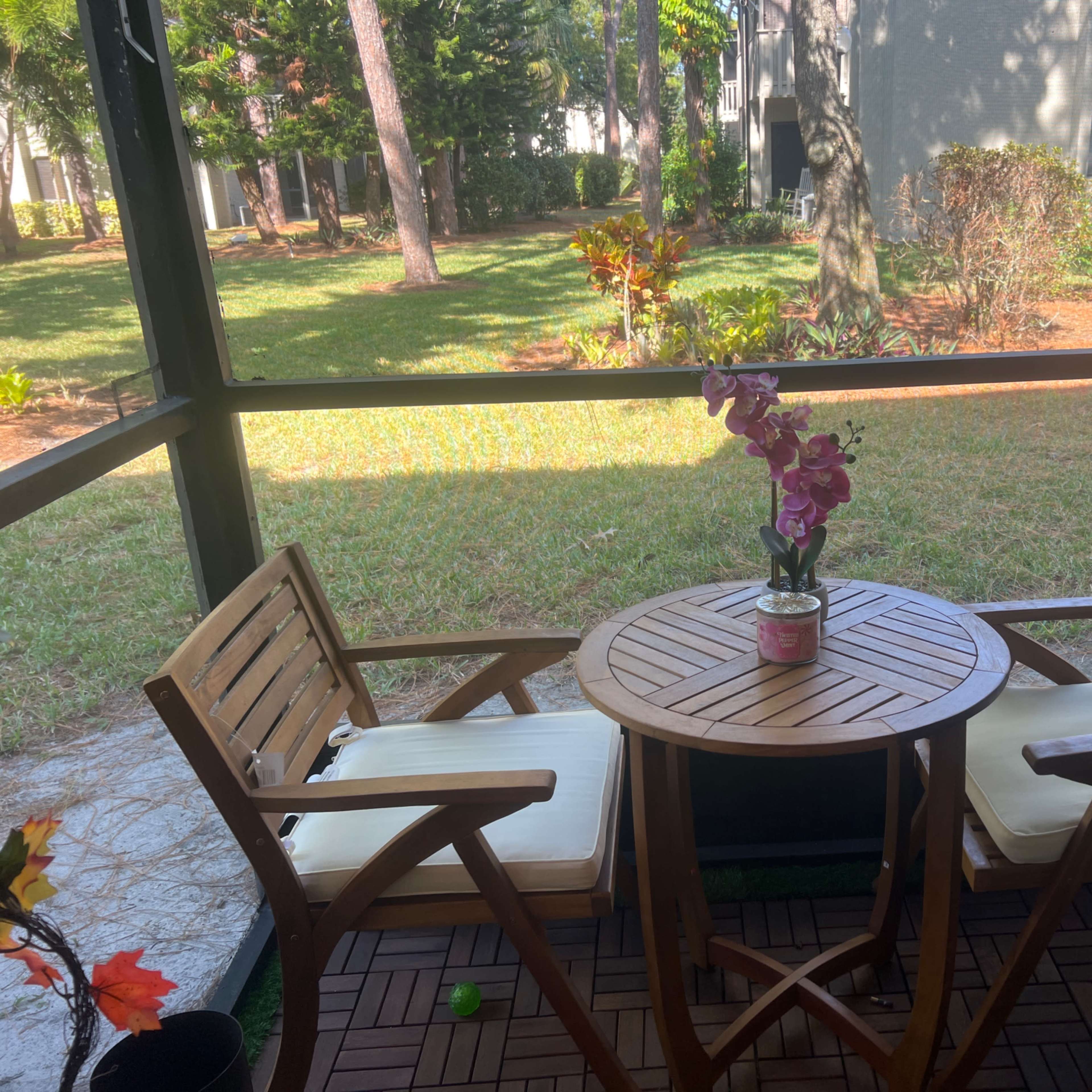 A small wooden table with two chairs and a vase of flowers sits on a screened porch, overlooking a grassy area and trees.