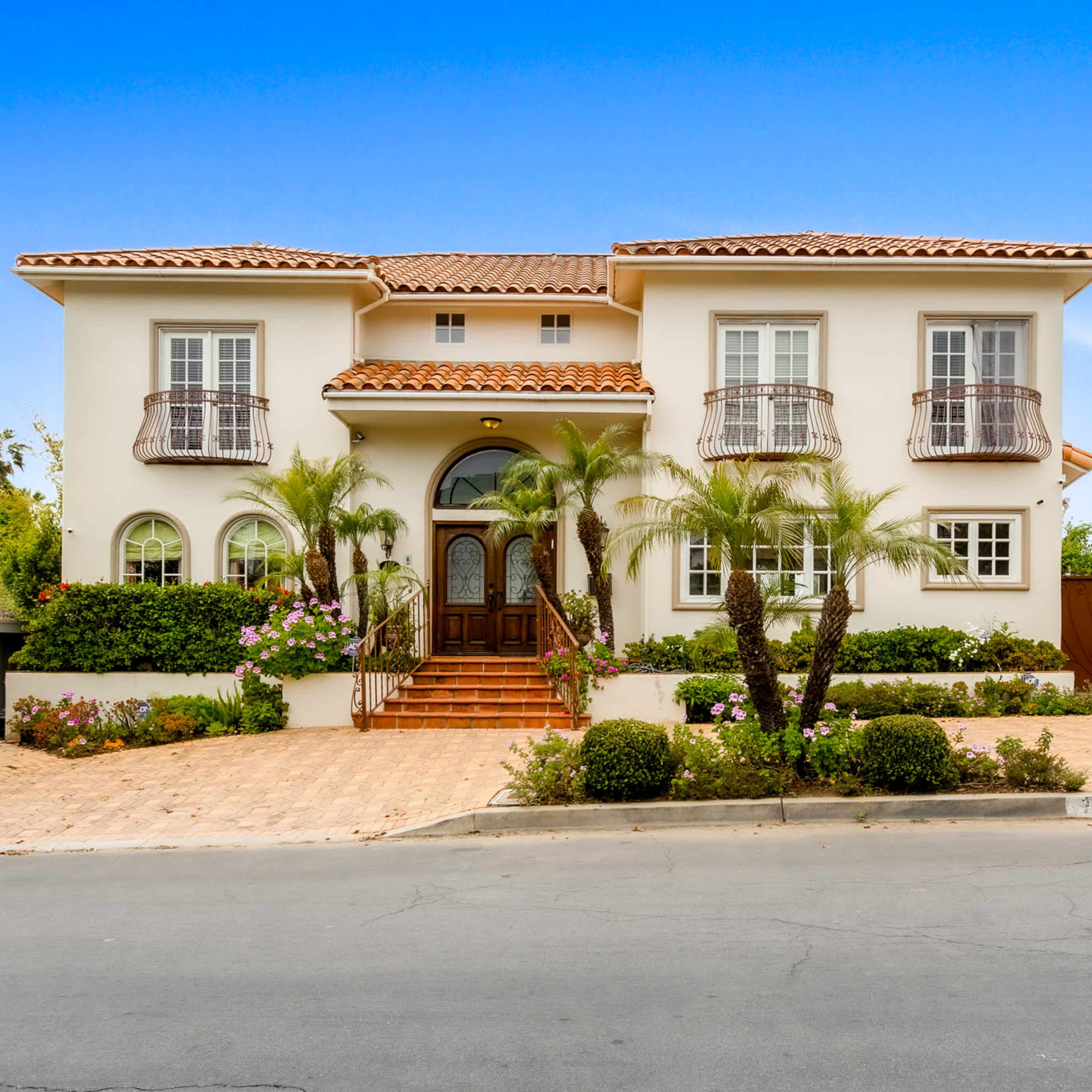 A two-story house with a tiled roof, large front windows, and palm trees is situated on a landscaped lot.
