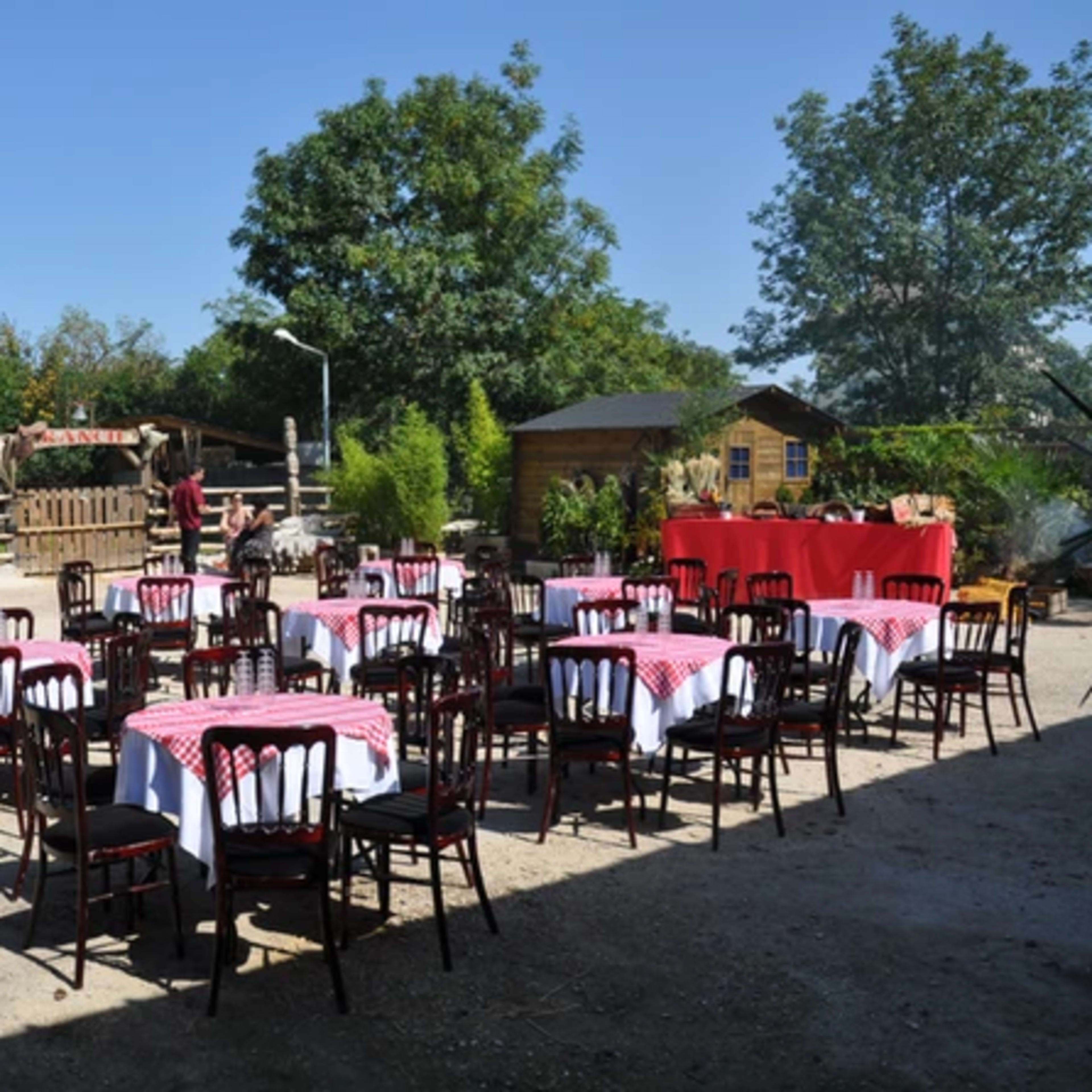 The scene shows an outdoor venue with several tables set up with checkered tablecloths amid greenery and structures, preparing for an event.
