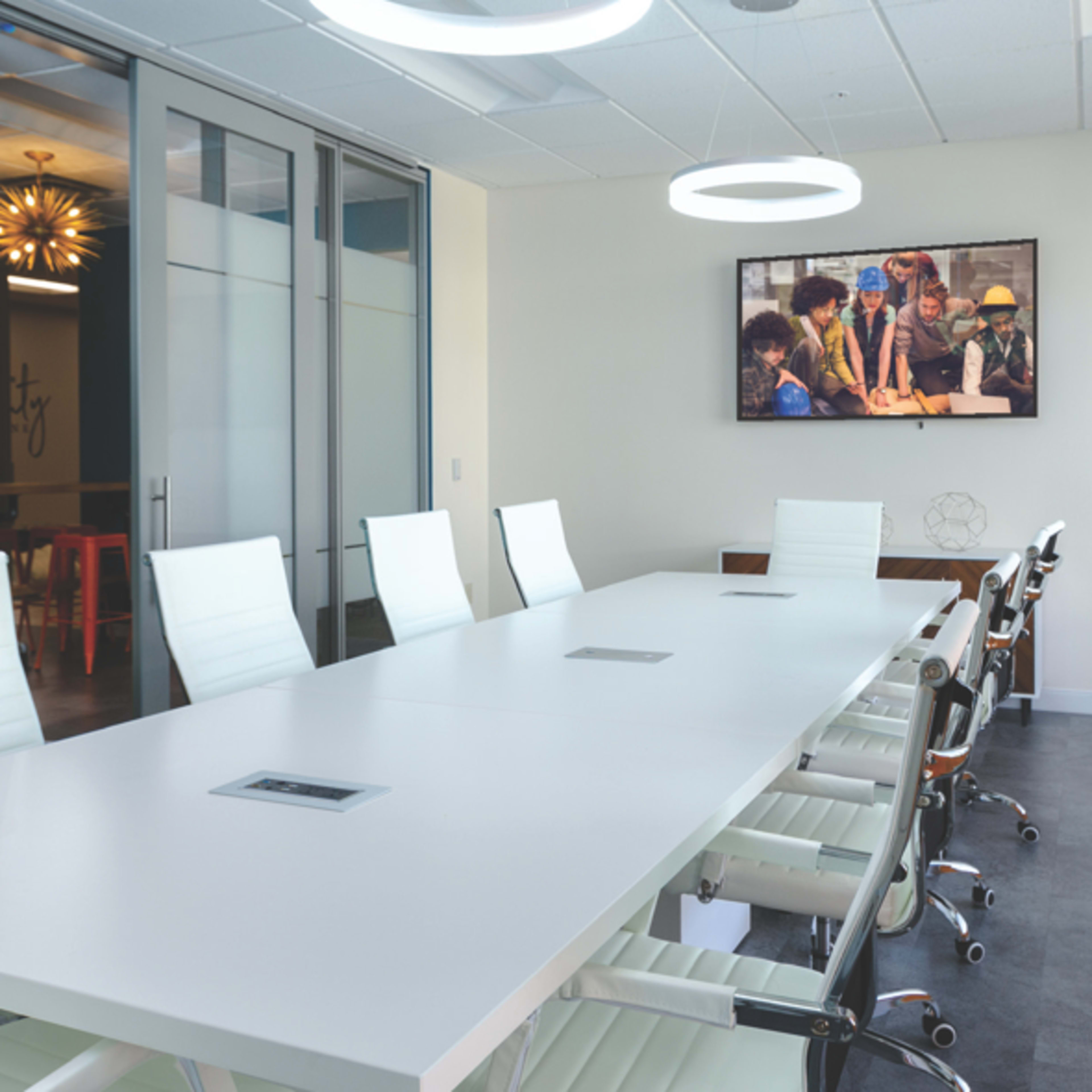 A modern conference room features a long white table surrounded by ergonomic chairs, with sliding glass doors and a television displaying a group of people collaborating.