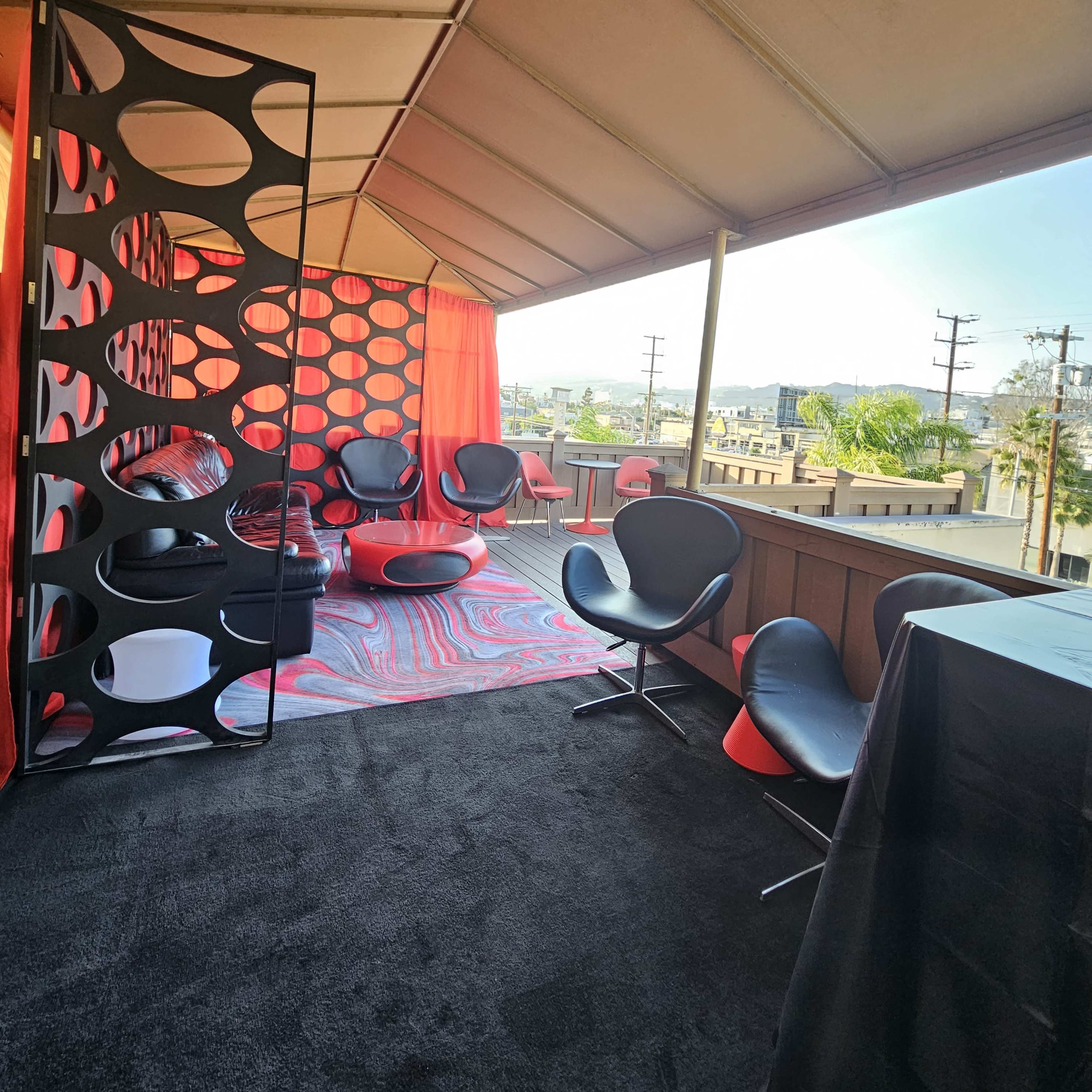 A modern rooftop lounge area with black and red seating, a patterned rug, and a view of the surrounding cityscape through an open canopy.