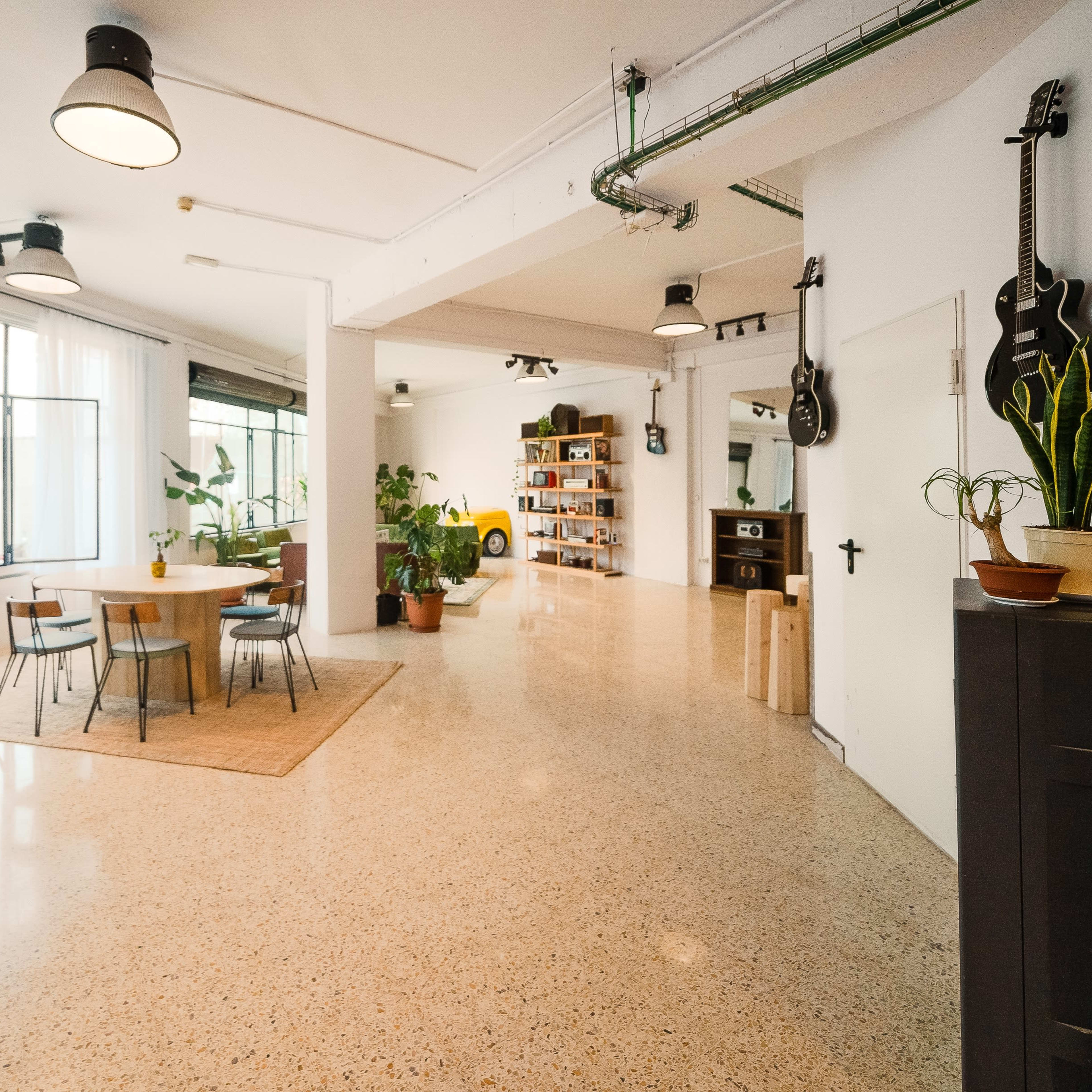 A spacious room with a dining table, plants, and a bookshelf, featuring large windows that allow natural light to enter.