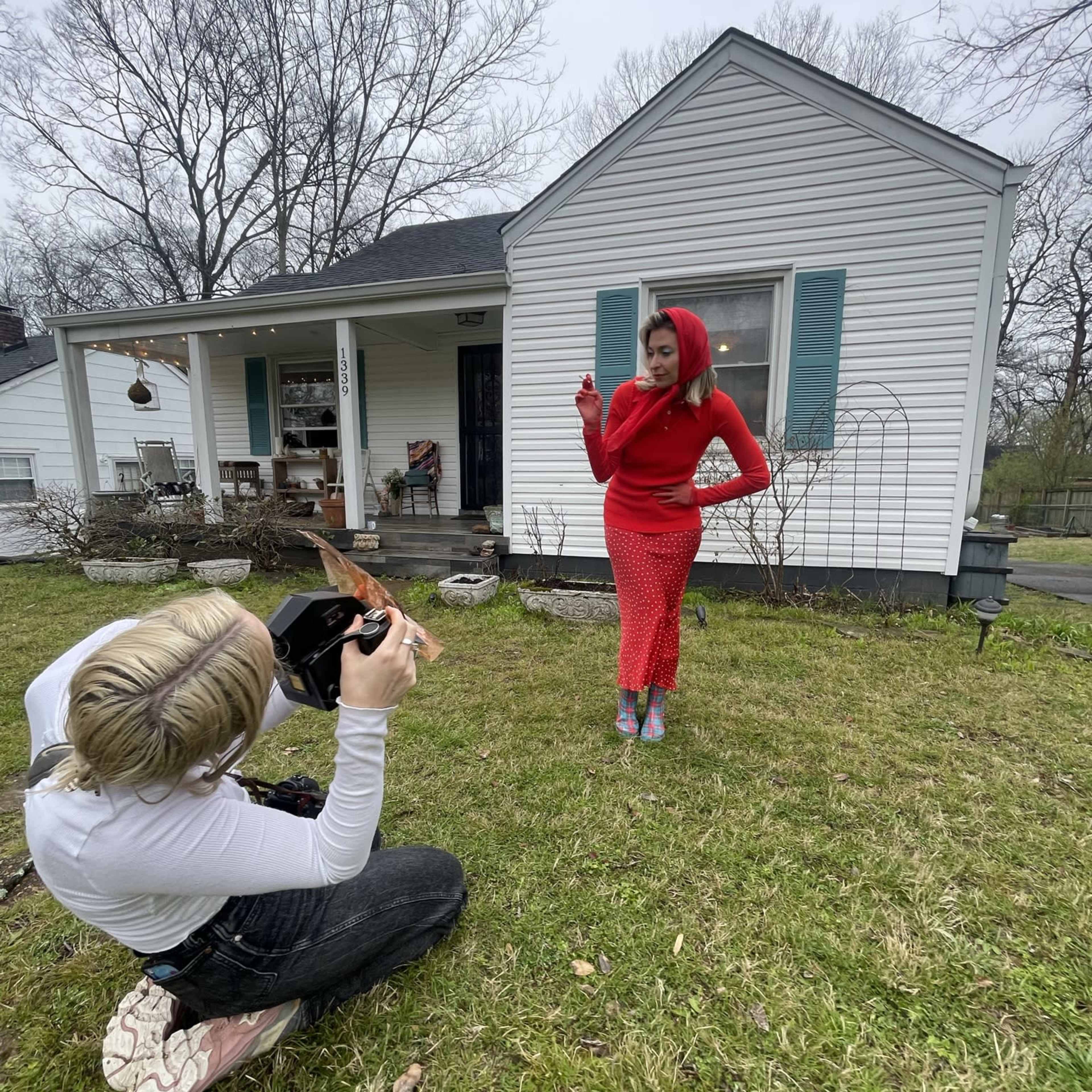 A photographer captures a model posing in a red outfit in front of a white house on a grassy lawn.