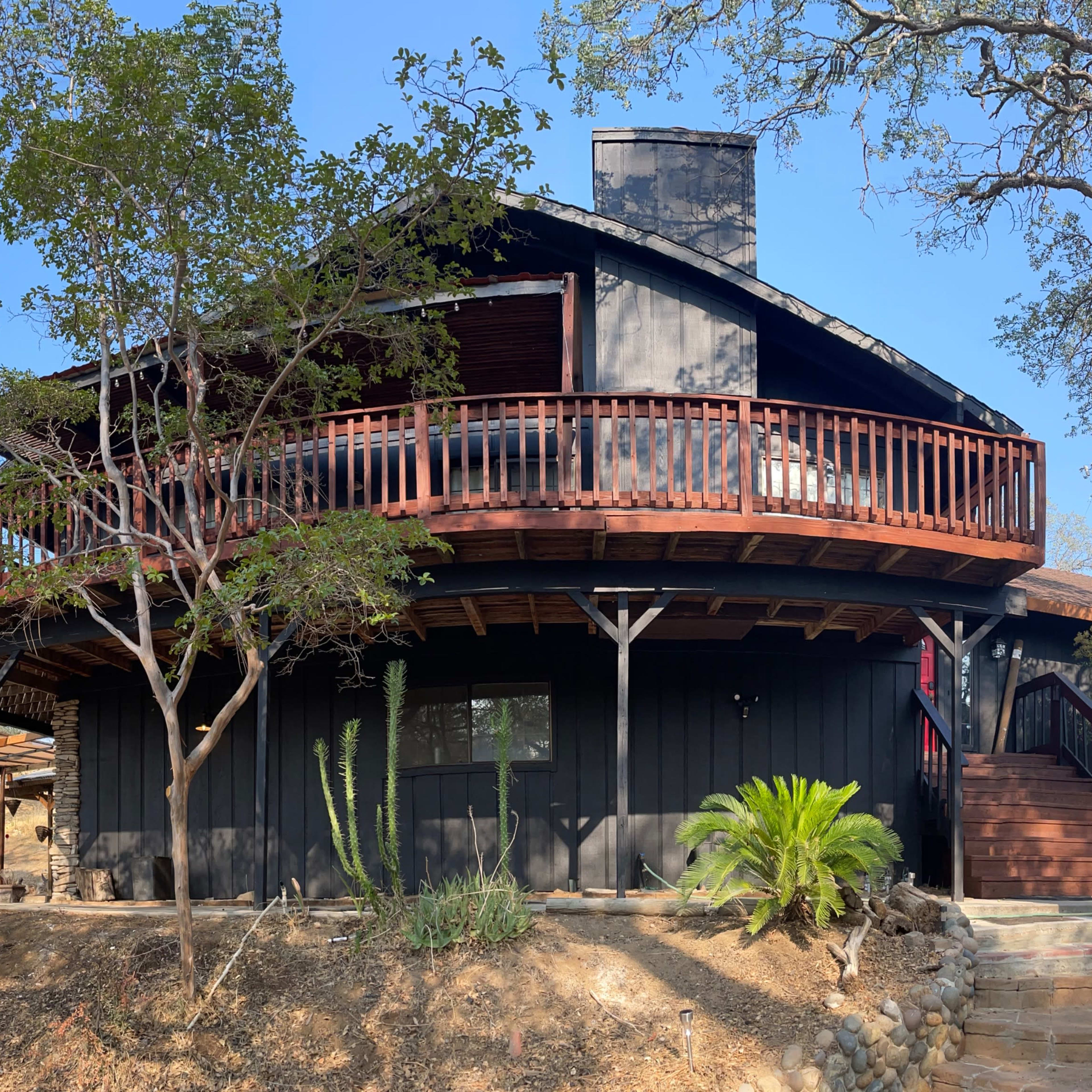 A modern, circular wooden house with a balcony, surrounded by trees and a landscaped area.