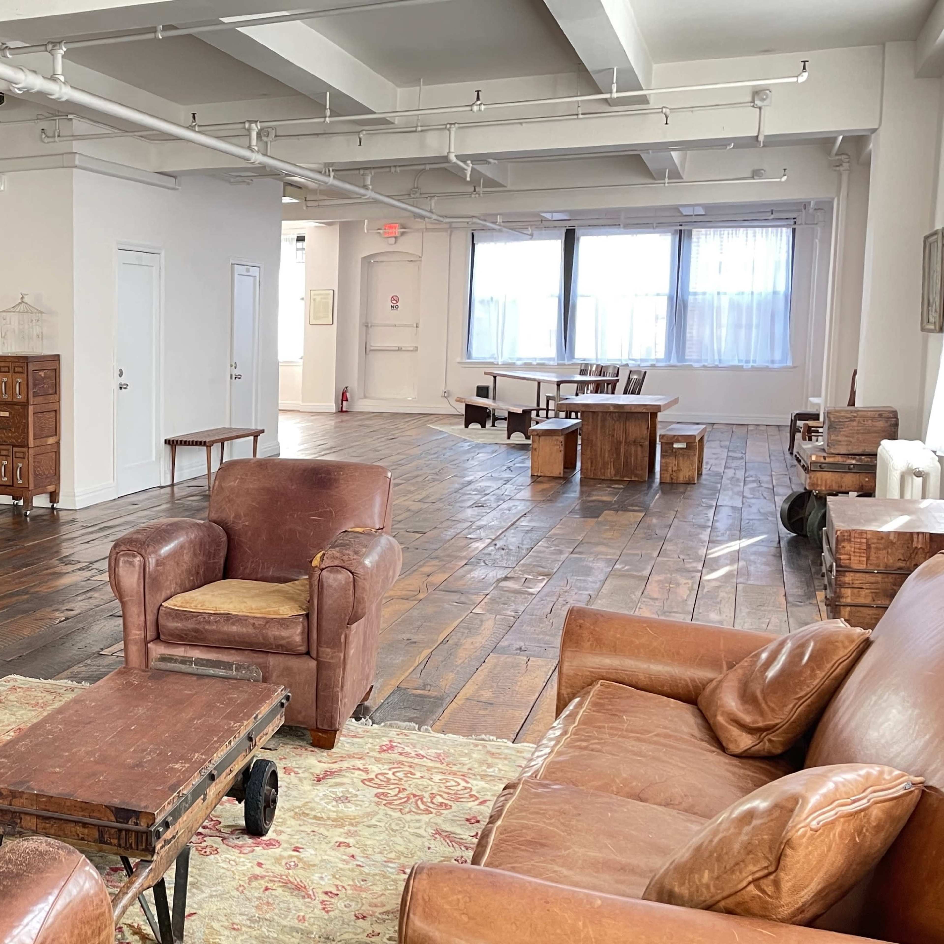 A spacious, well-lit room with wooden floors, featuring a seating area with brown leather chairs, a coffee table, and various wooden furniture pieces along the walls.