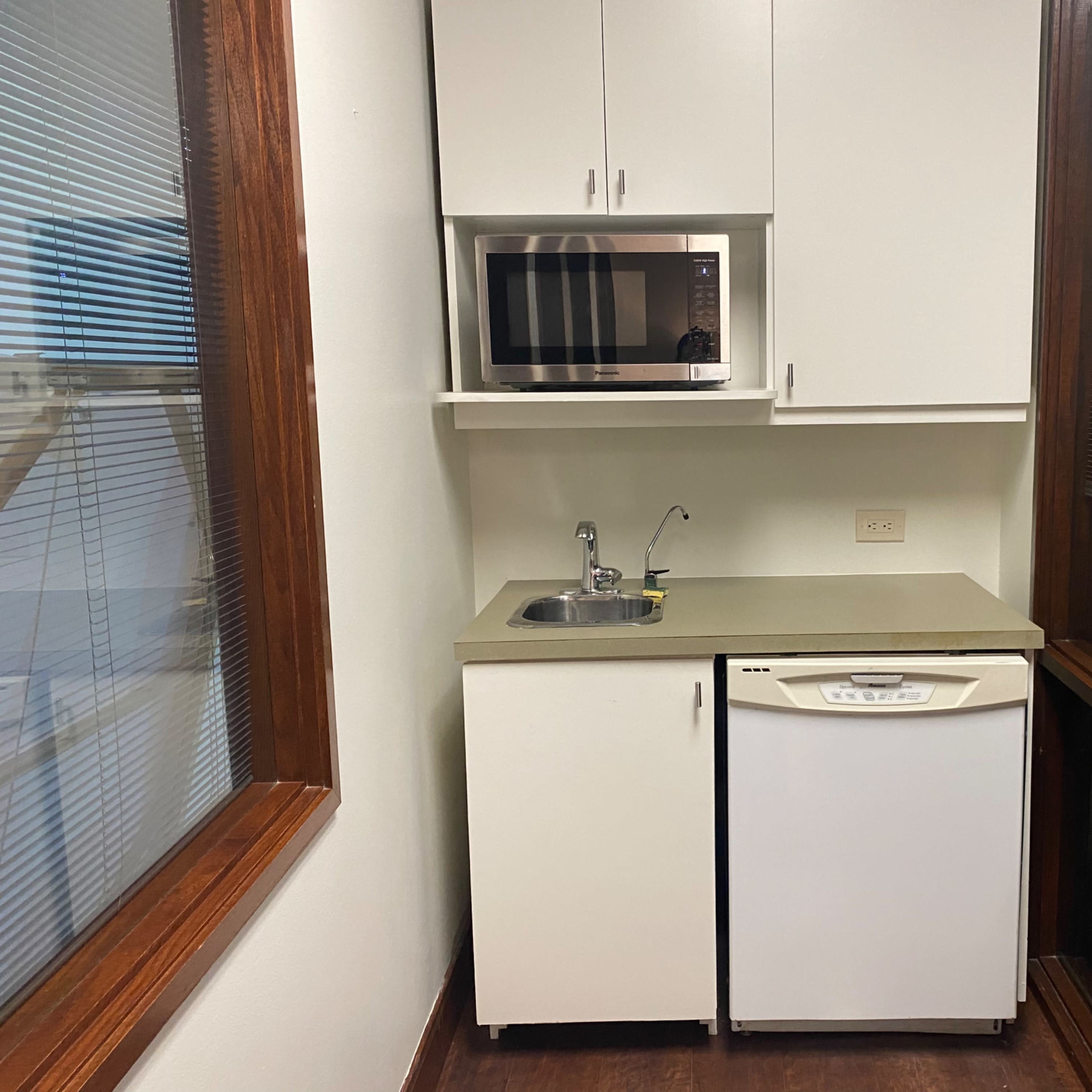 A small kitchen area with a microwave, sink, and refrigerator against a wall with windows.