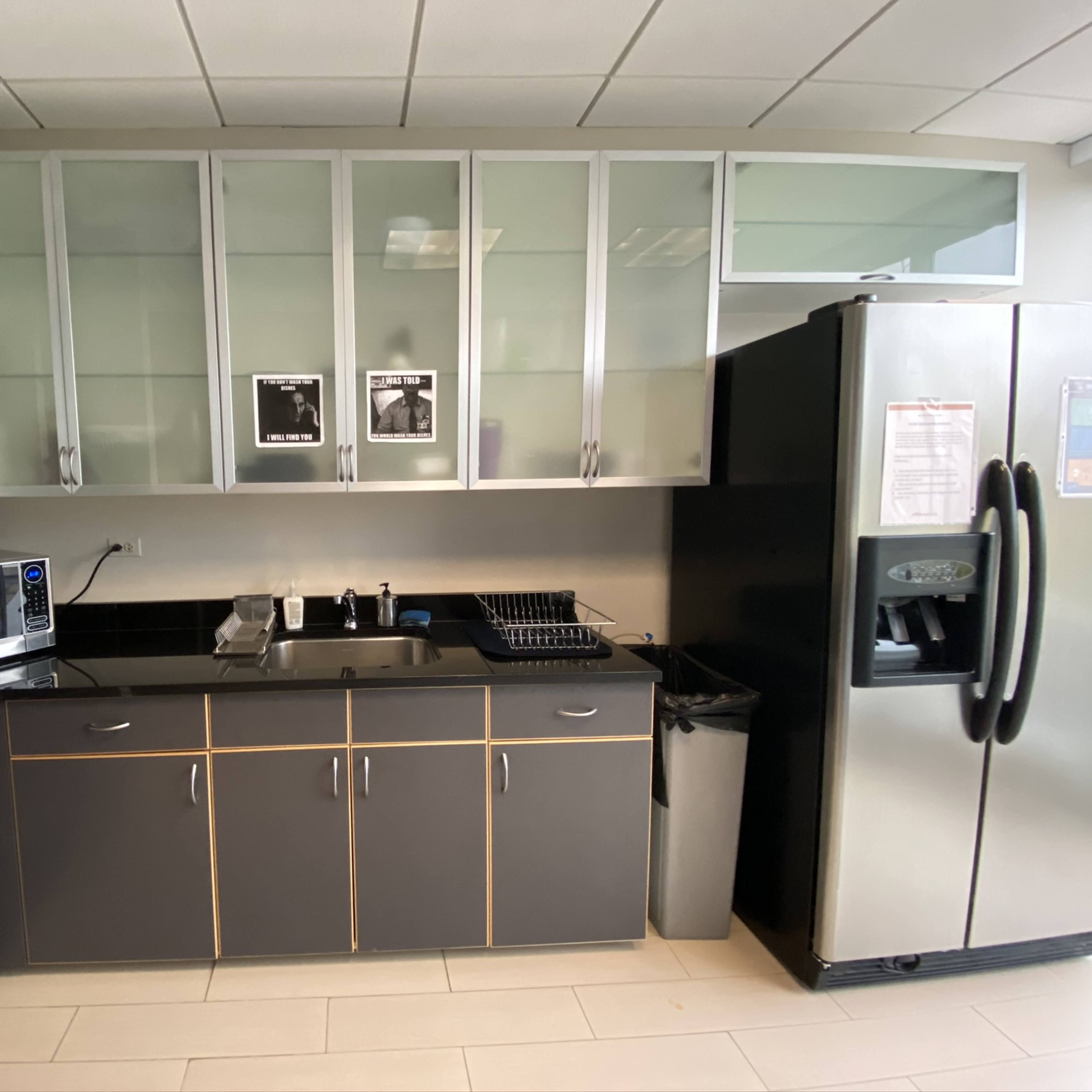 A modern kitchen with gray cabinets, a stainless steel refrigerator, a microwave, and a coffee maker, along with a dish rack and a trash bin on a black countertop.