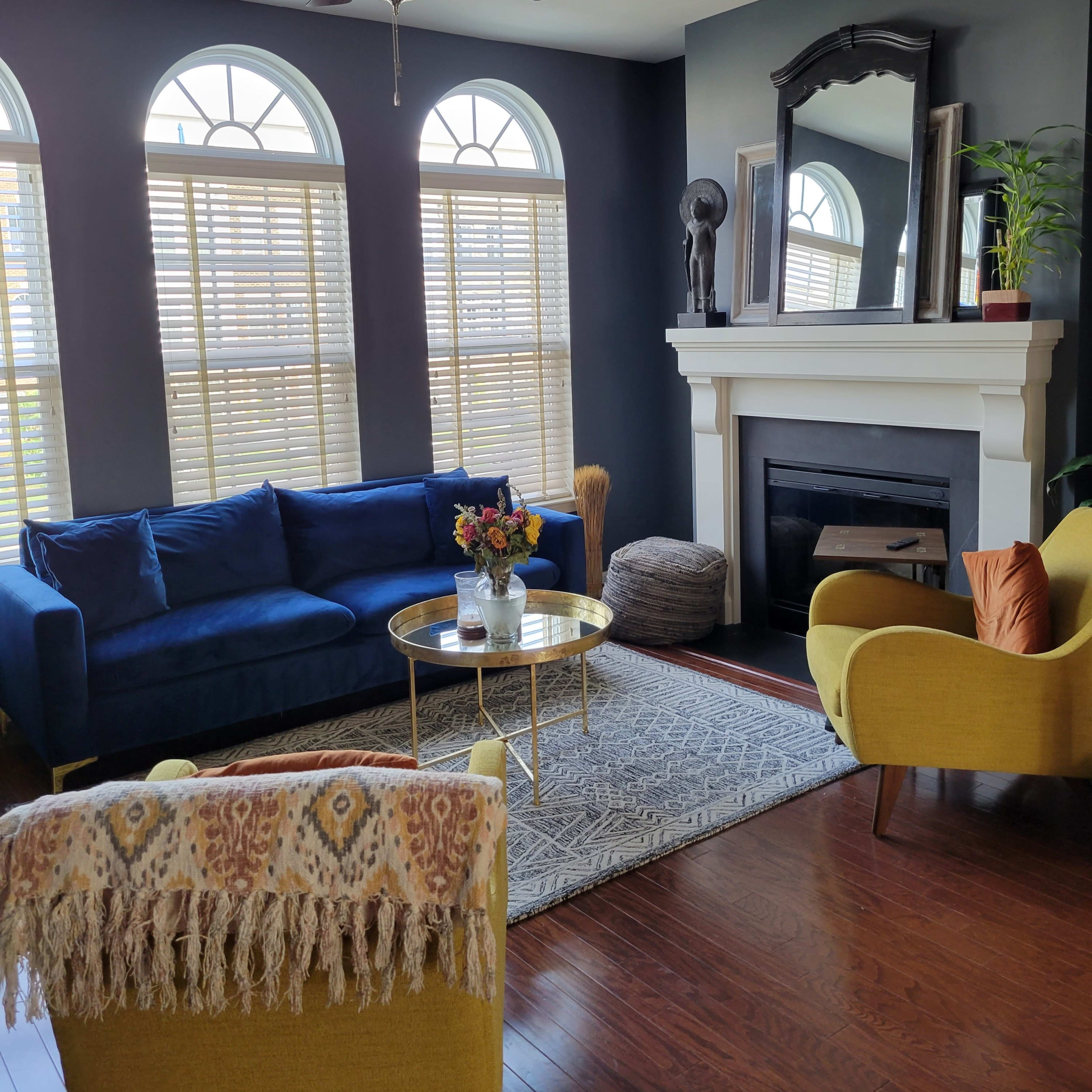 A living room features a blue sofa, yellow chairs, a round coffee table, and a fireplace beneath a large mirror, with natural light streaming through the arched windows.