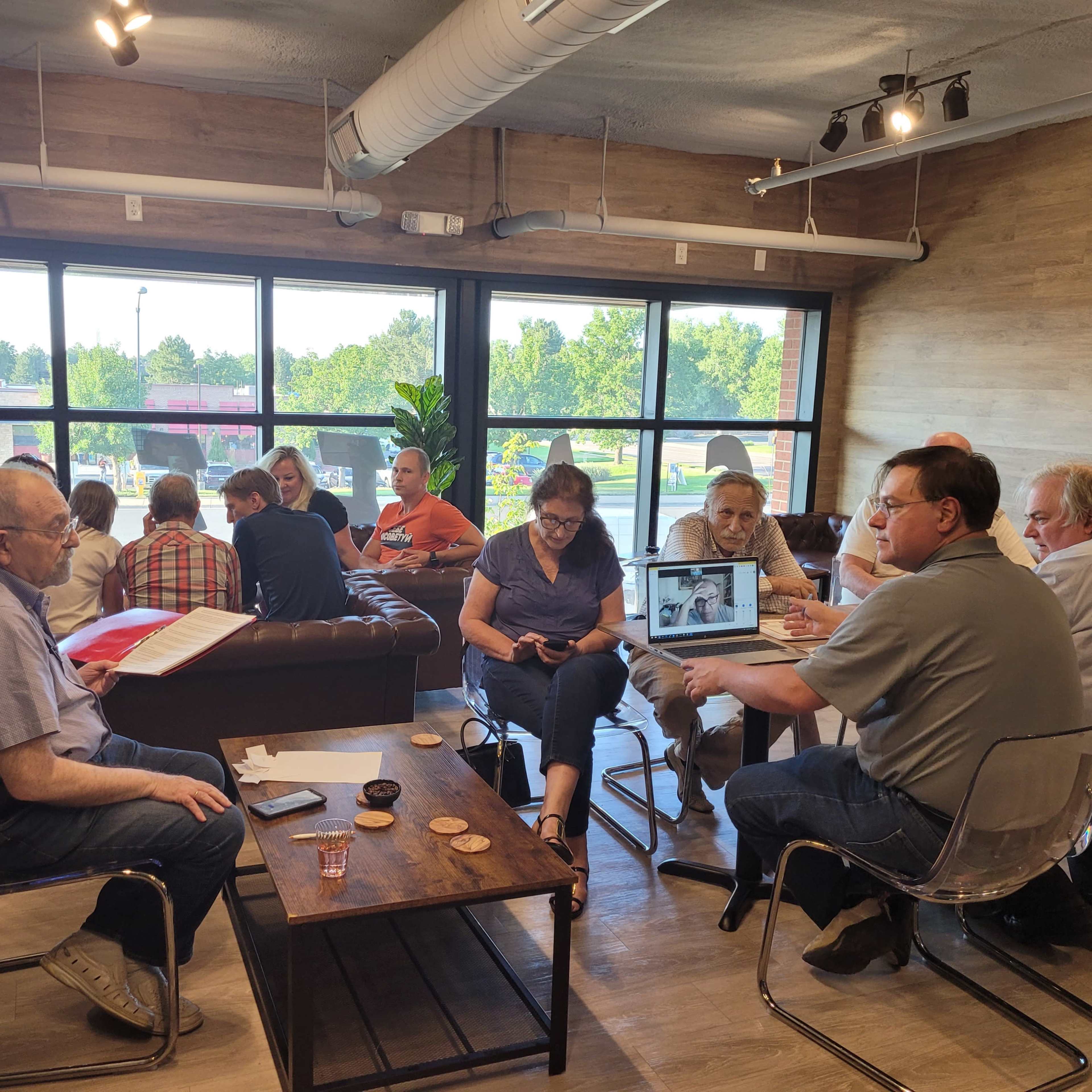 A group of people is gathered in a well-lit café, engaged in conversation and using laptops around wooden tables.
