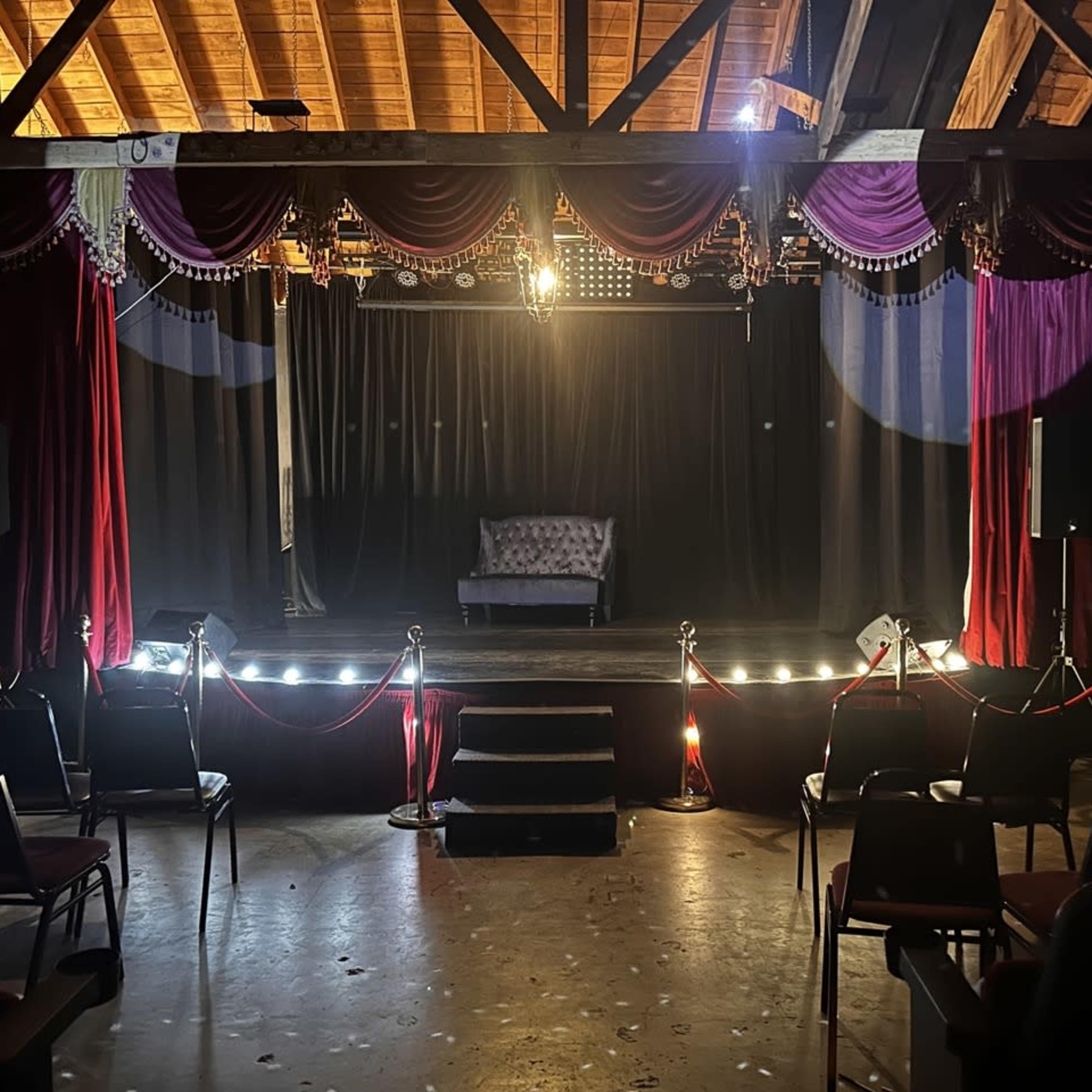 A small stage is set with a lone couch and illuminated by spotlights, surrounded by empty chairs and red velvet curtains.