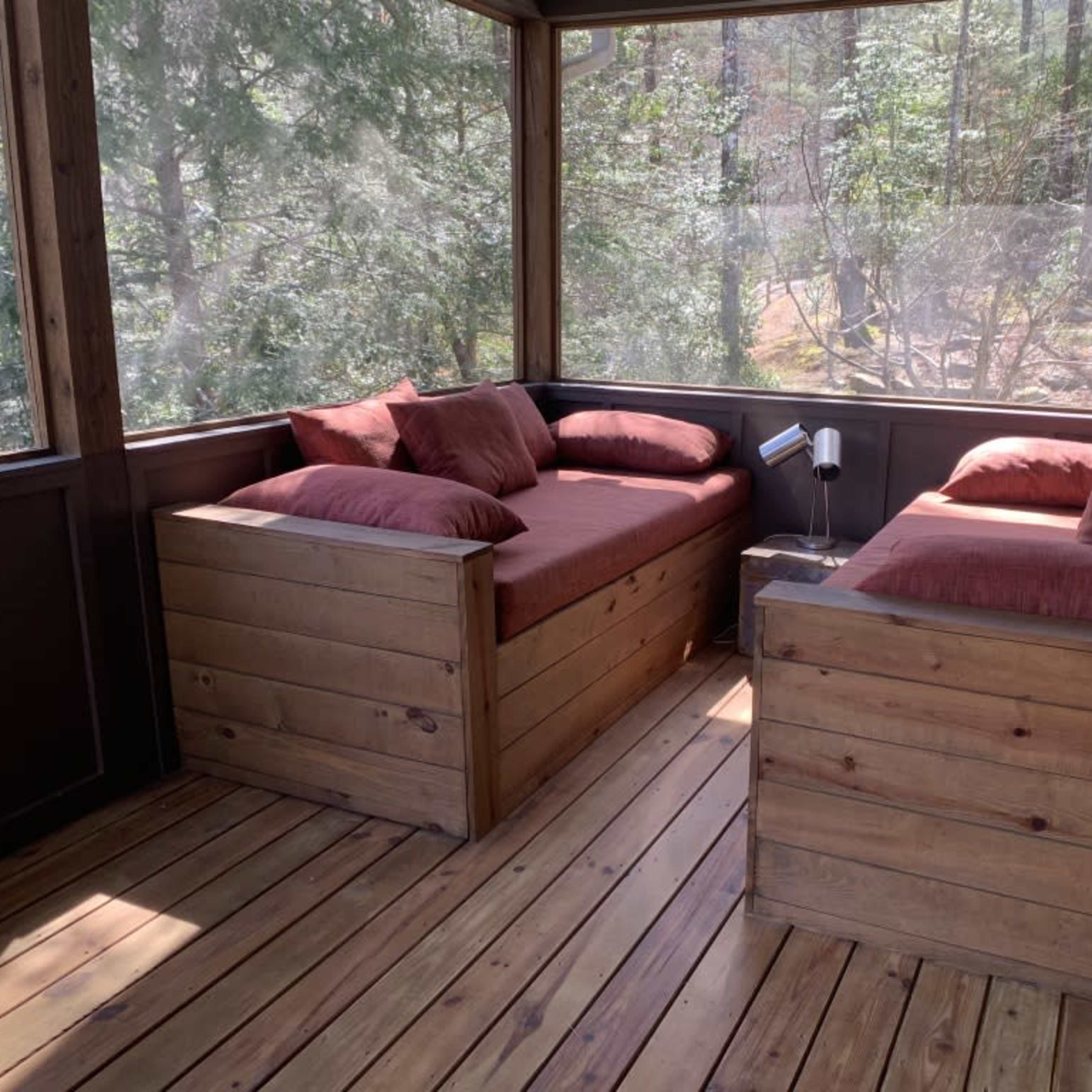 A cozy screened-in porch with two wooden seating areas filled with red cushions, surrounded by green trees.