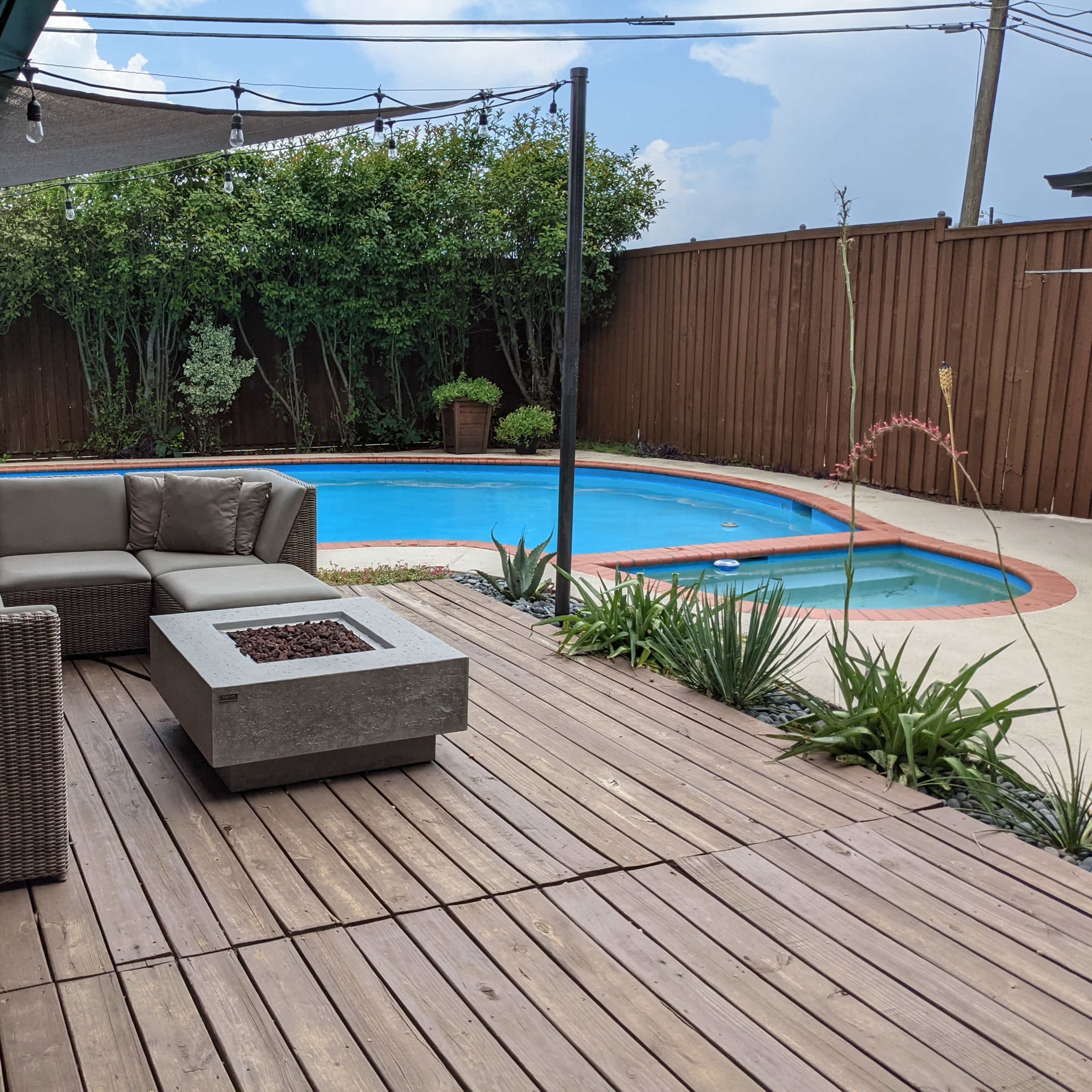 A wooden deck featuring a seating area with a sectional sofa and a fire pit overlooks a swimming pool surrounded by landscaping.